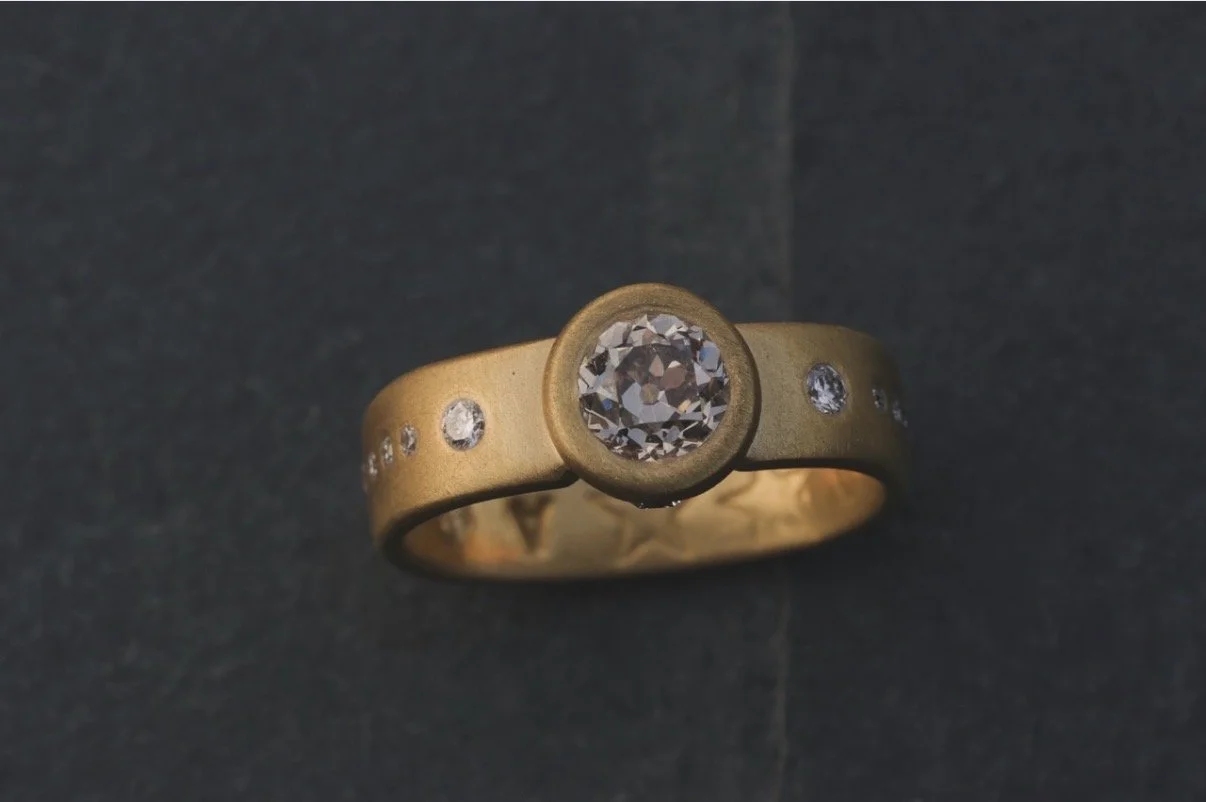 Gold ring with a central round diamond and smaller diamonds on the band, placed on a dark surface.