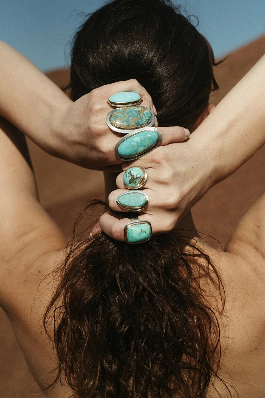 A person with long brown hair, sitting with arms raised behind their head, wearing multiple large turquoise rings on fingers, showing off their hair.