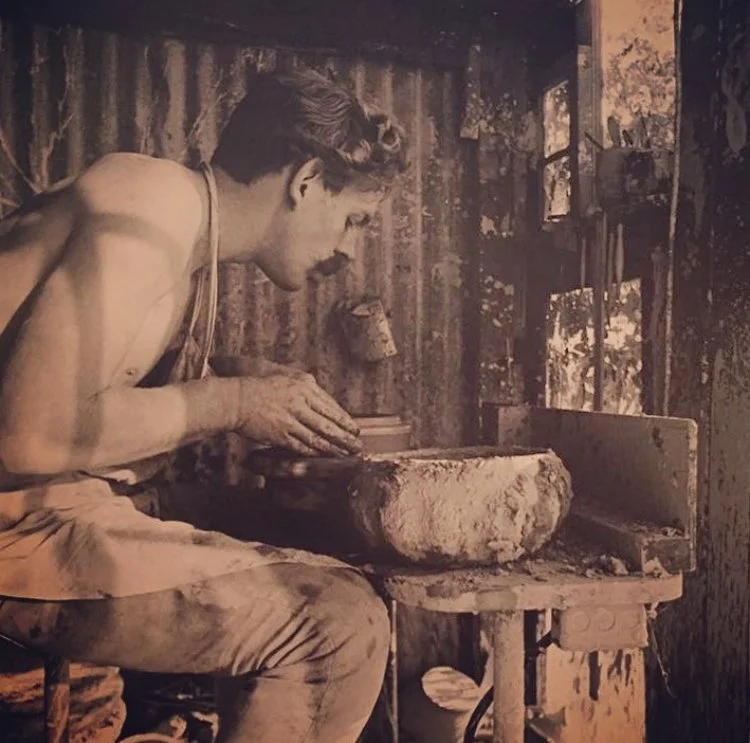 A young man sculpting clay on a pottery wheel inside a rustic workshop, with wooden walls and natural light coming through a window.