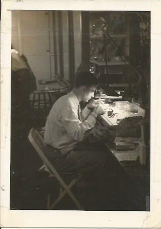 A man sitting at a desk, looking through a microscope in a room filled with people and equipment.