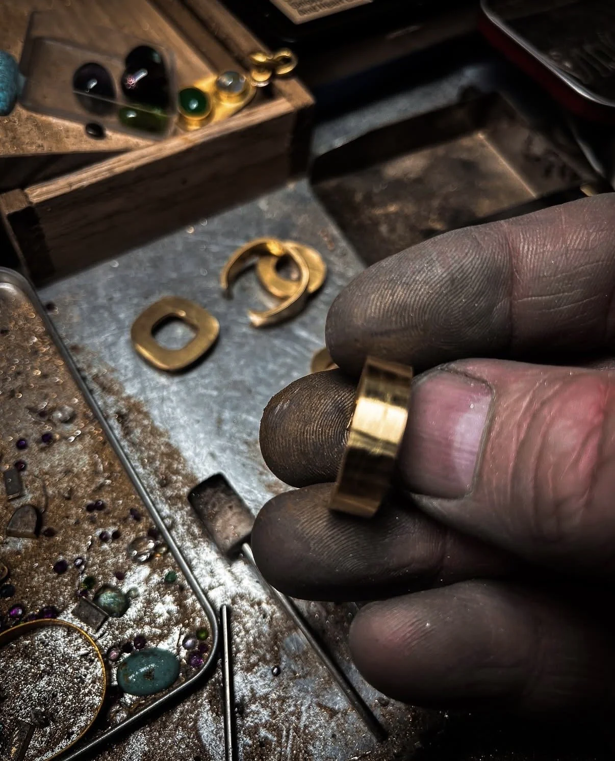 Close-up of a craftsman's hand holding a gold ring, with a jewelry-making workspace in the background showing tools, small beads, and metal pieces.