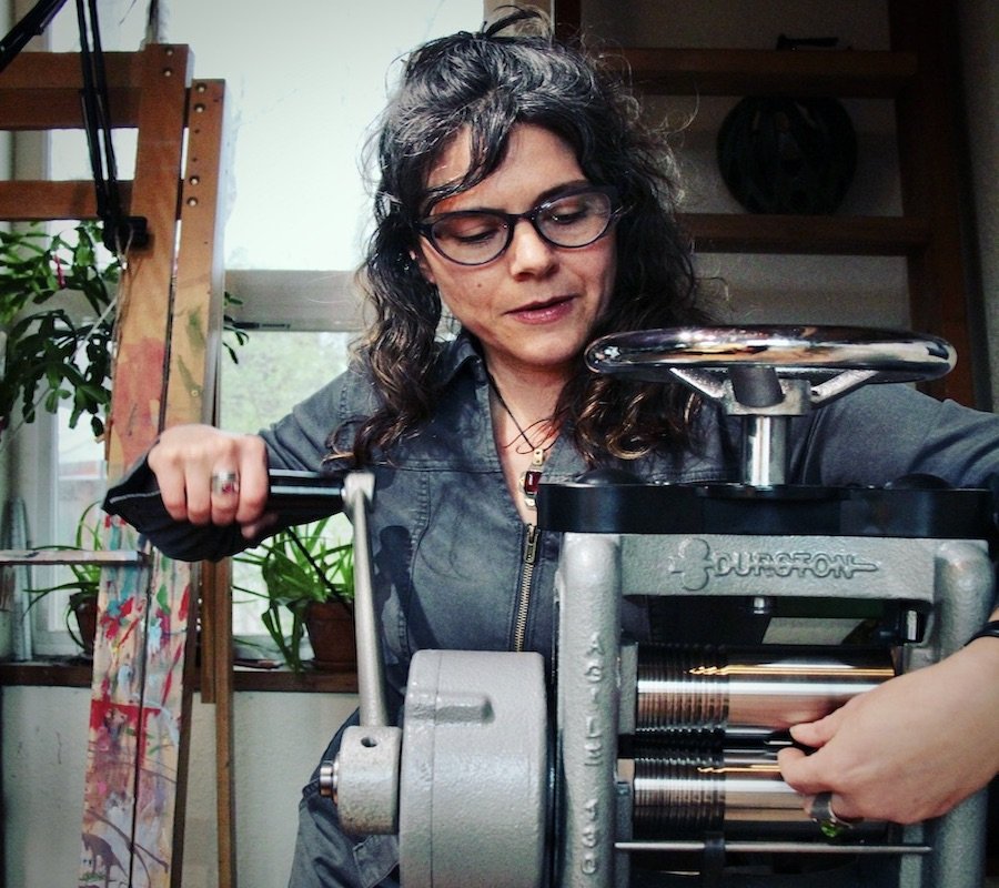 Woman wearing glasses using a vintage hand-crank coffee roaster in a cozy, plant-filled room.