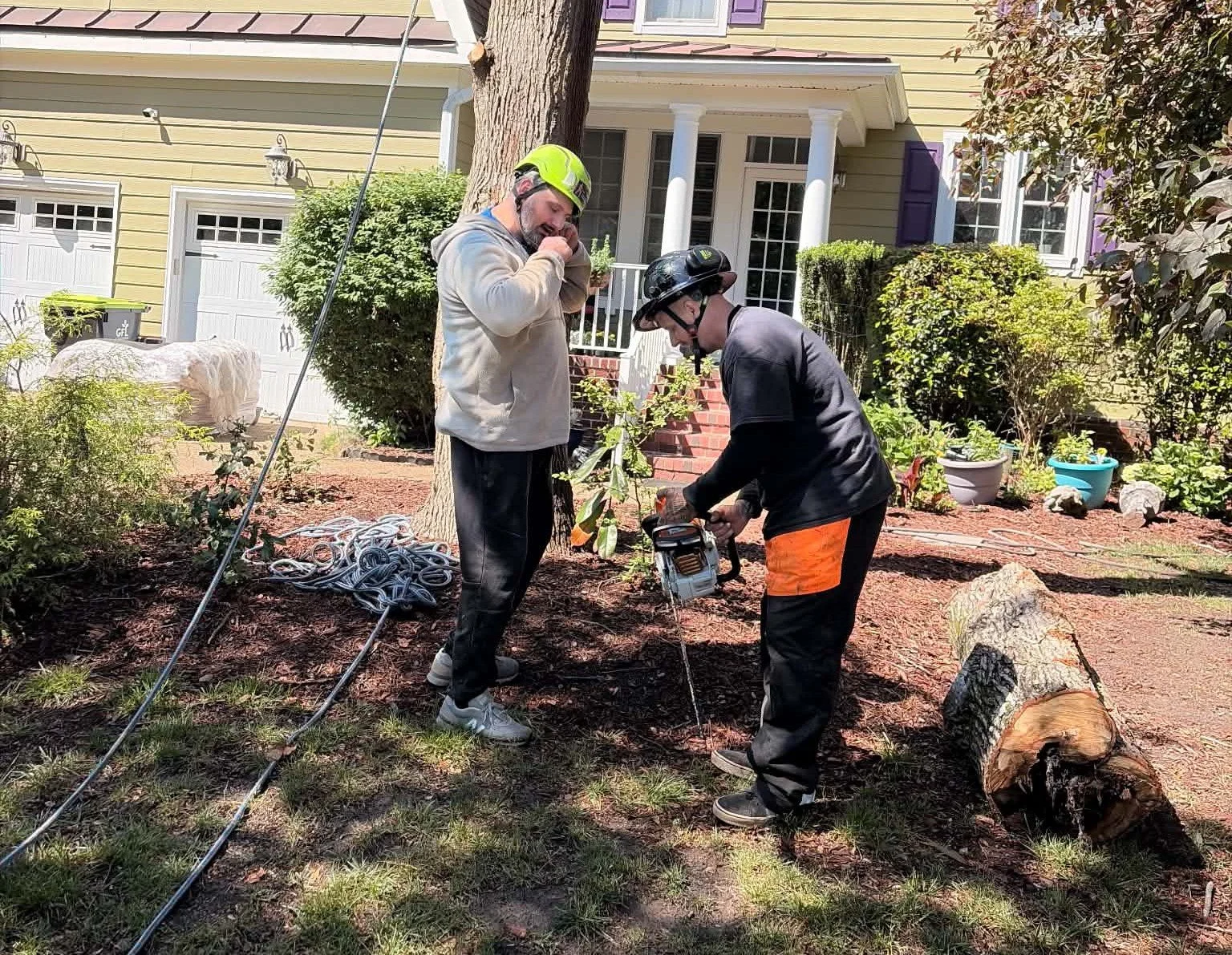 Two men wearing safety helmets and work clothes are cutting a fallen tree trunk with a chainsaw while removing a tree in Virginia Beach, VA