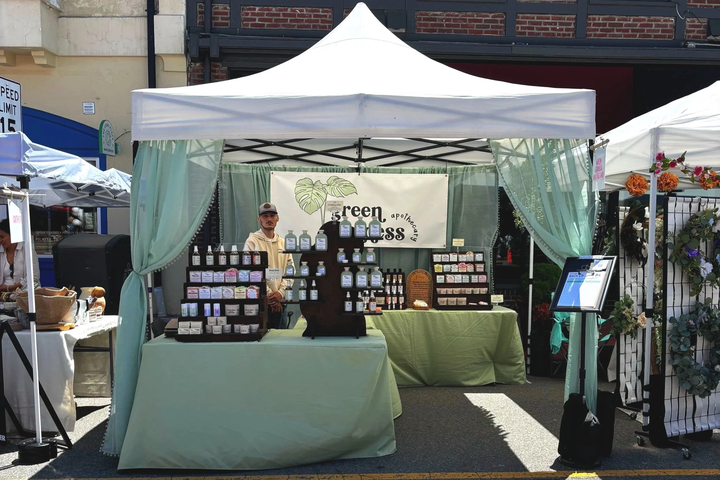 Outdoor market stall with a white canopy selling various bottles and jars, with a man standing behind the display. Surrounding stalls and a brick building are visible in the background.