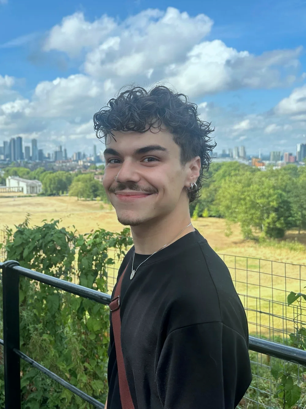 Smiling young man with curly hair and earrings standing outdoors on a sunny day, with a city skyline and green park in the background.