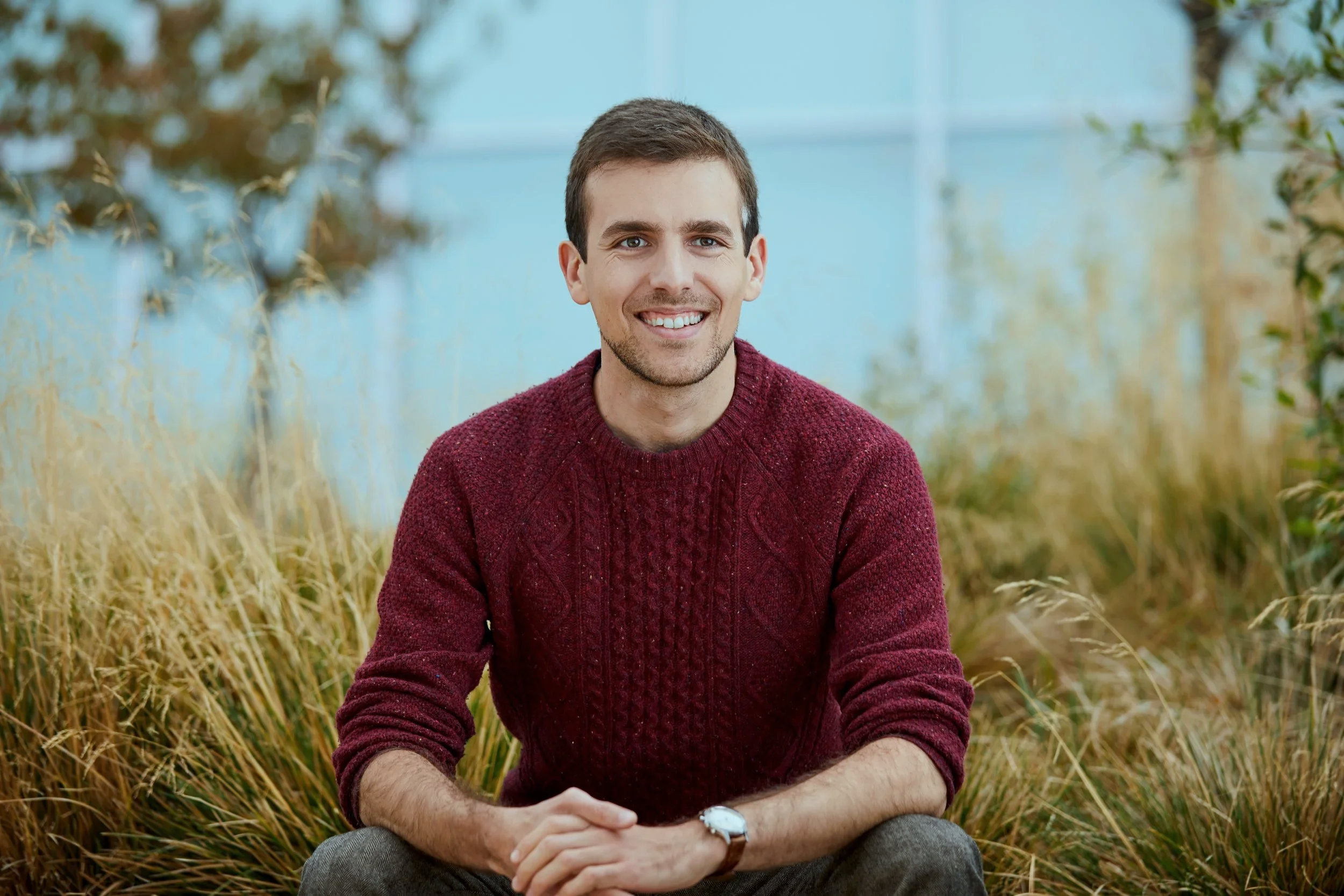 A smiling man with short brown hair, wearing a maroon sweater, sitting outdoors among tall grass.
