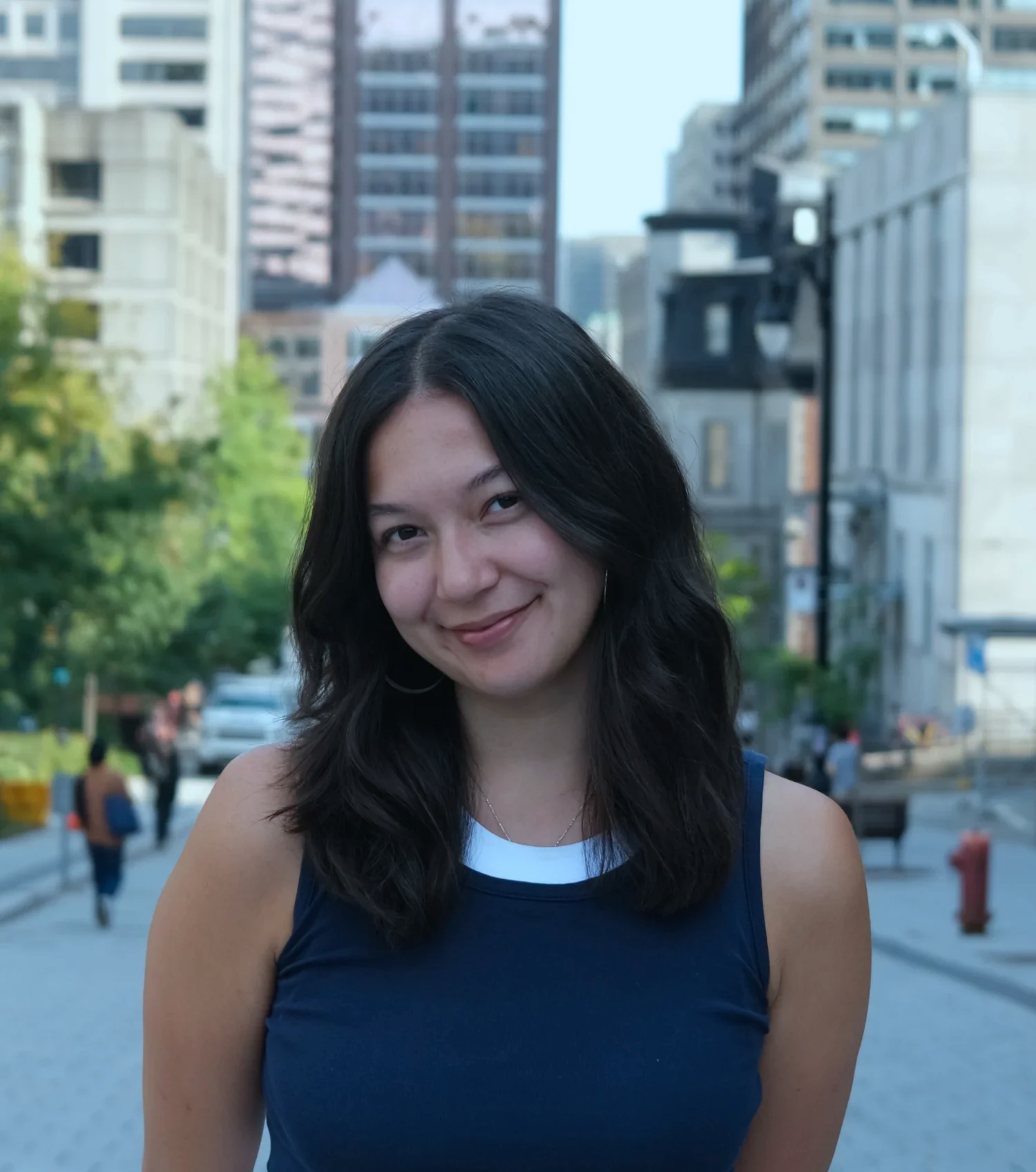 Young woman with dark hair smiling outdoors in an urban setting with tall buildings and trees in the background.