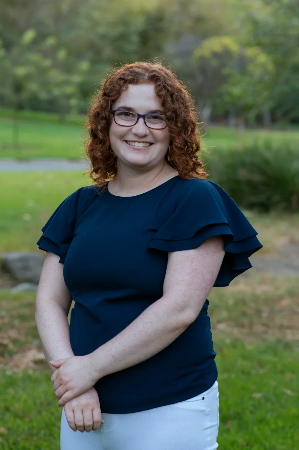 Woman smiling outdoors with green trees and grass in the background, wearing glasses, a navy blue ruffled sleeve top, and white pants.