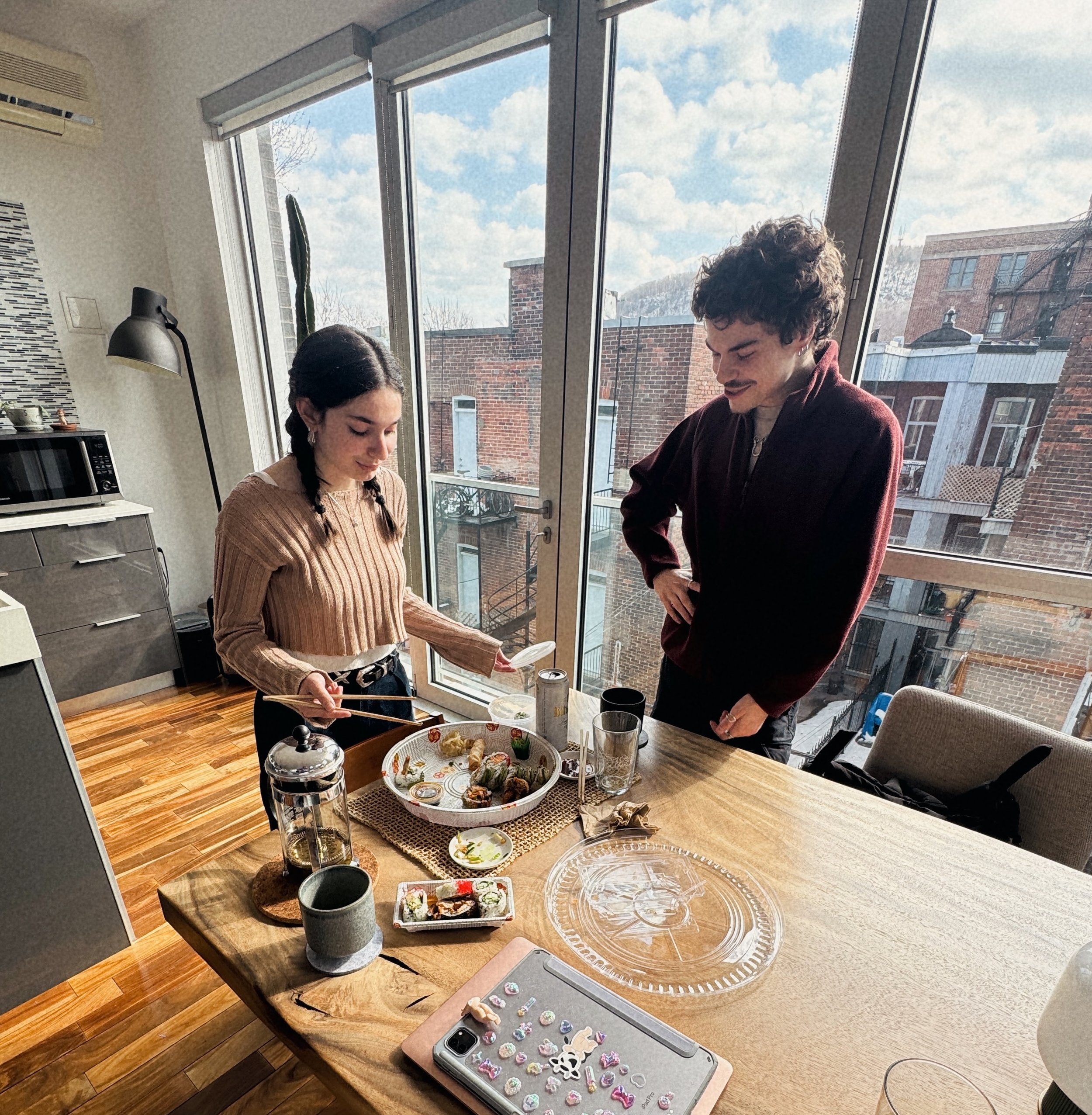 Two people standing at a kitchen table with sushi on a large platter and various drink glasses. The scene is lit by natural daylight coming through large windows.