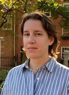 A person with shoulder-length brown hair wearing a blue and white striped shirt, standing outside with trees and a brick building in the background.