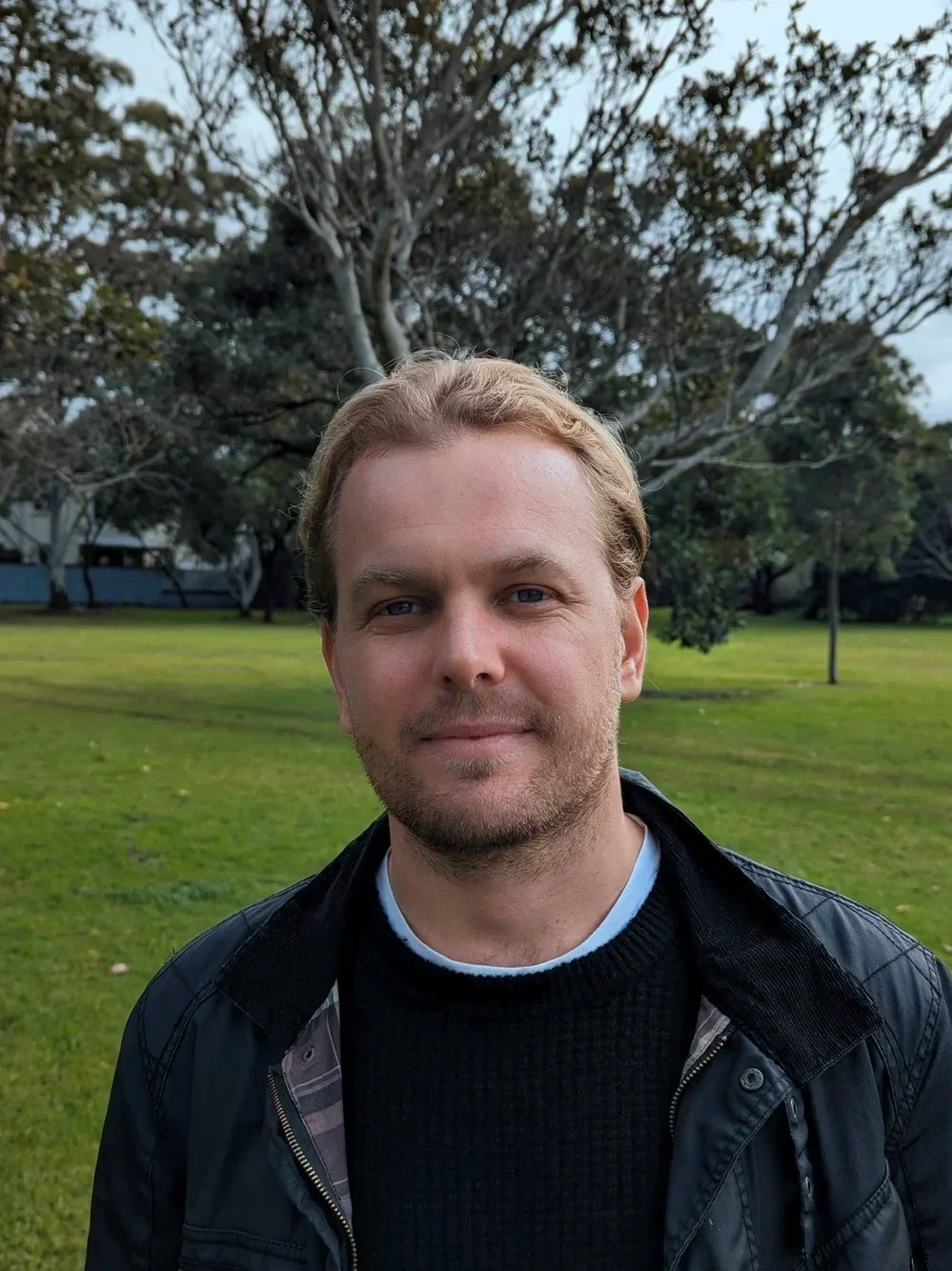 A man with blond hair and a beard standing outdoors in a park with green grass and trees in the background.