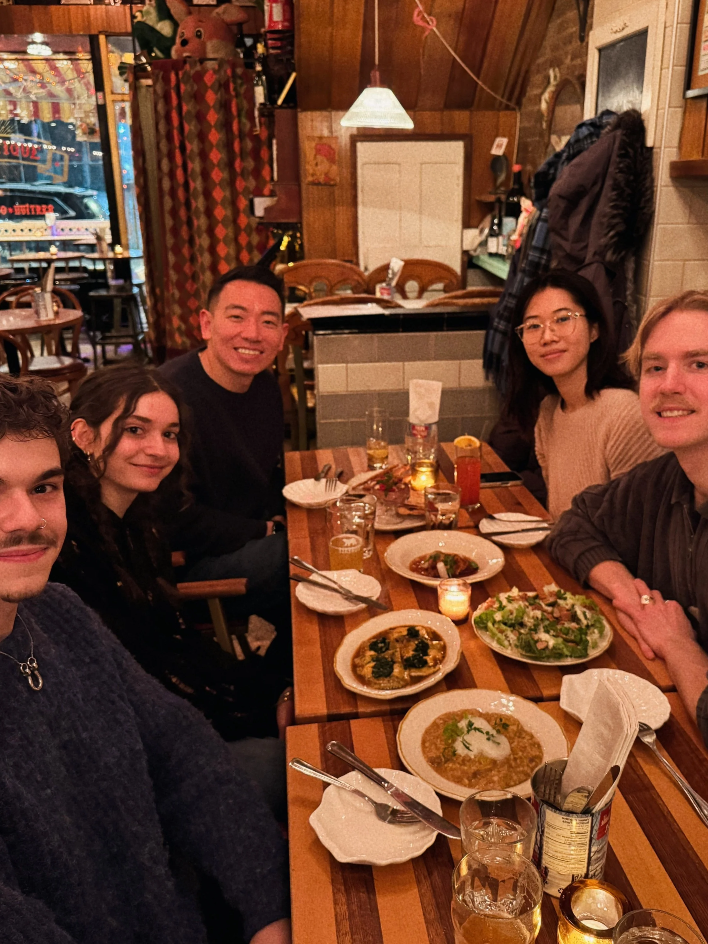 A group of six people are gathered around a wooden table in a cozy restaurant. The table is filled with plates of food, glasses of drinks, and utensils. The restaurant has warm lighting, wooden walls, and a window showing the street outside with some visible signs.