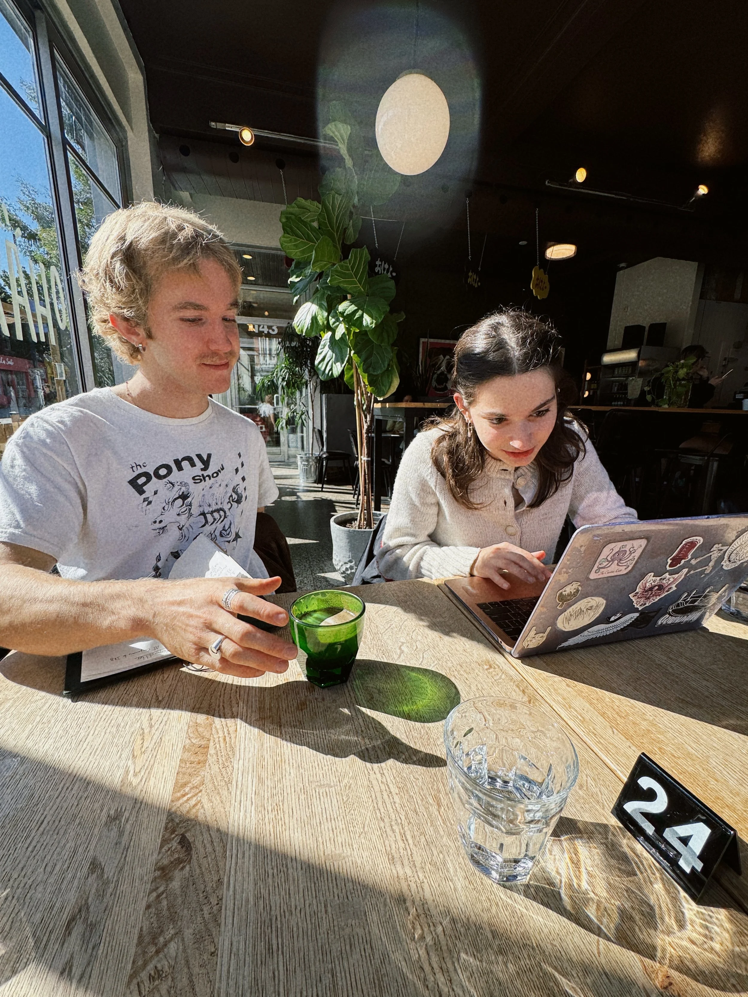 Two people sitting at a wooden table in a cafe. The person on the left has light curly hair, is wearing a white t-shirt, and is holding a green glass. The person on the right has dark wavy hair, is wearing a cream-colored sweater, and is using a laptop decorated with stickers. There is a glass of water and a table marker with the number 24 on the table. The cafe has large windows, plants, and warm lighting.