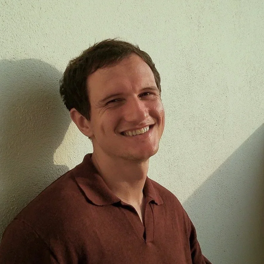 A smiling man with short dark hair wearing a brown polo shirt, standing against a plain light-colored wall.