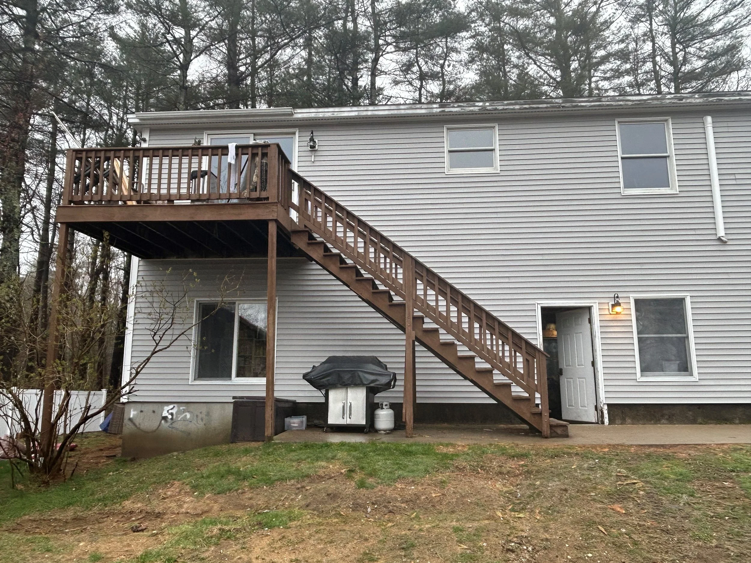 Back of a house with a wooden deck and staircase, a grill, and a propane tank; trees in the background.