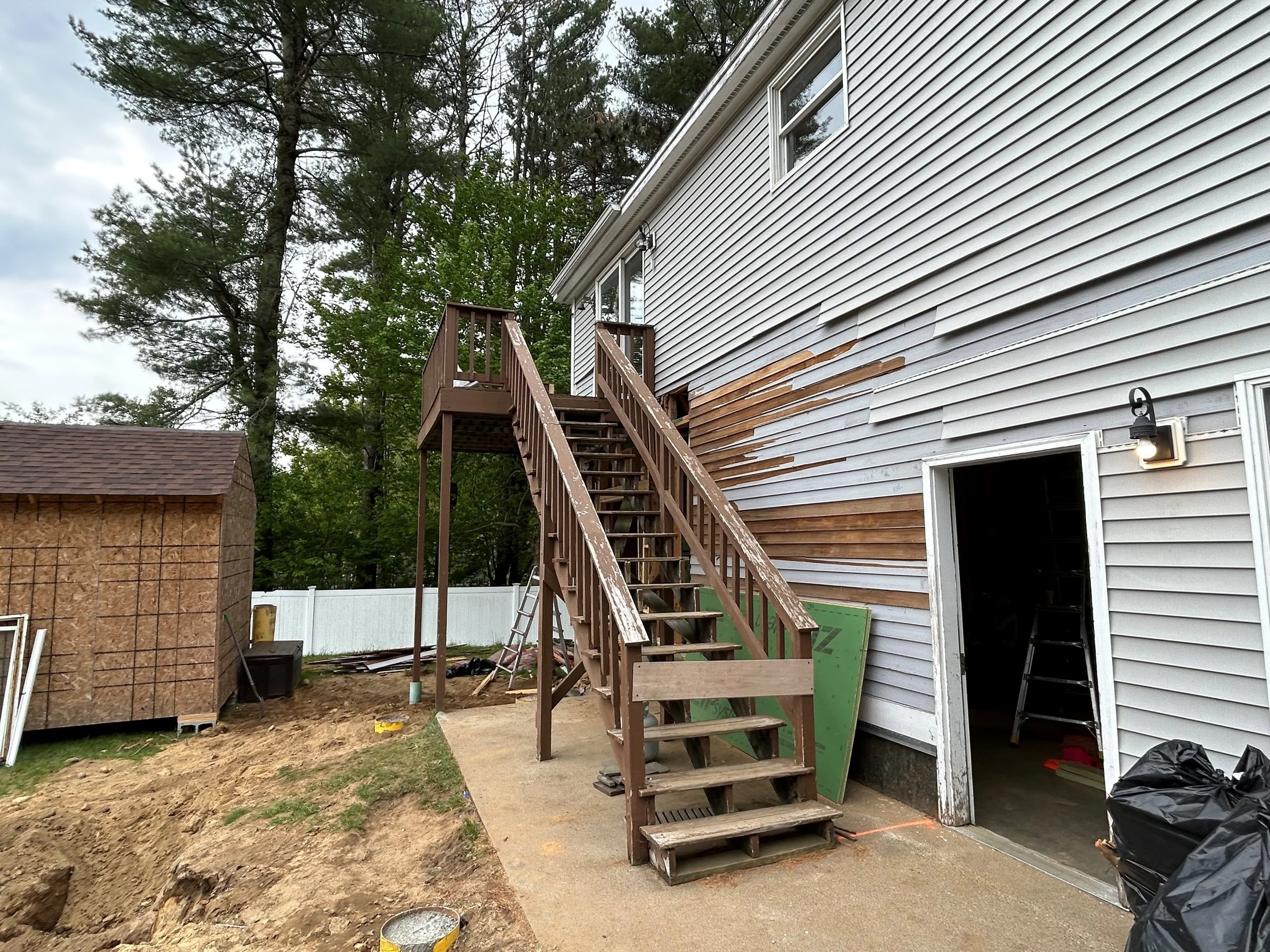 Backyard construction site with a wooden staircase attached to the side of a house that has grey siding, some of which has been recently replaced. There is a small shed on the left, and the ground is uneven with construction materials and tools scatt
