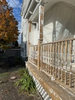 Side view of a house porch with a wooden railing, some peeling paint, and a plastic covering on part of the porch. There are trees with autumn foliage in the background.