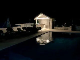 Nighttime scene of a swimming pool with a small pavilion and lounge chairs nearby.