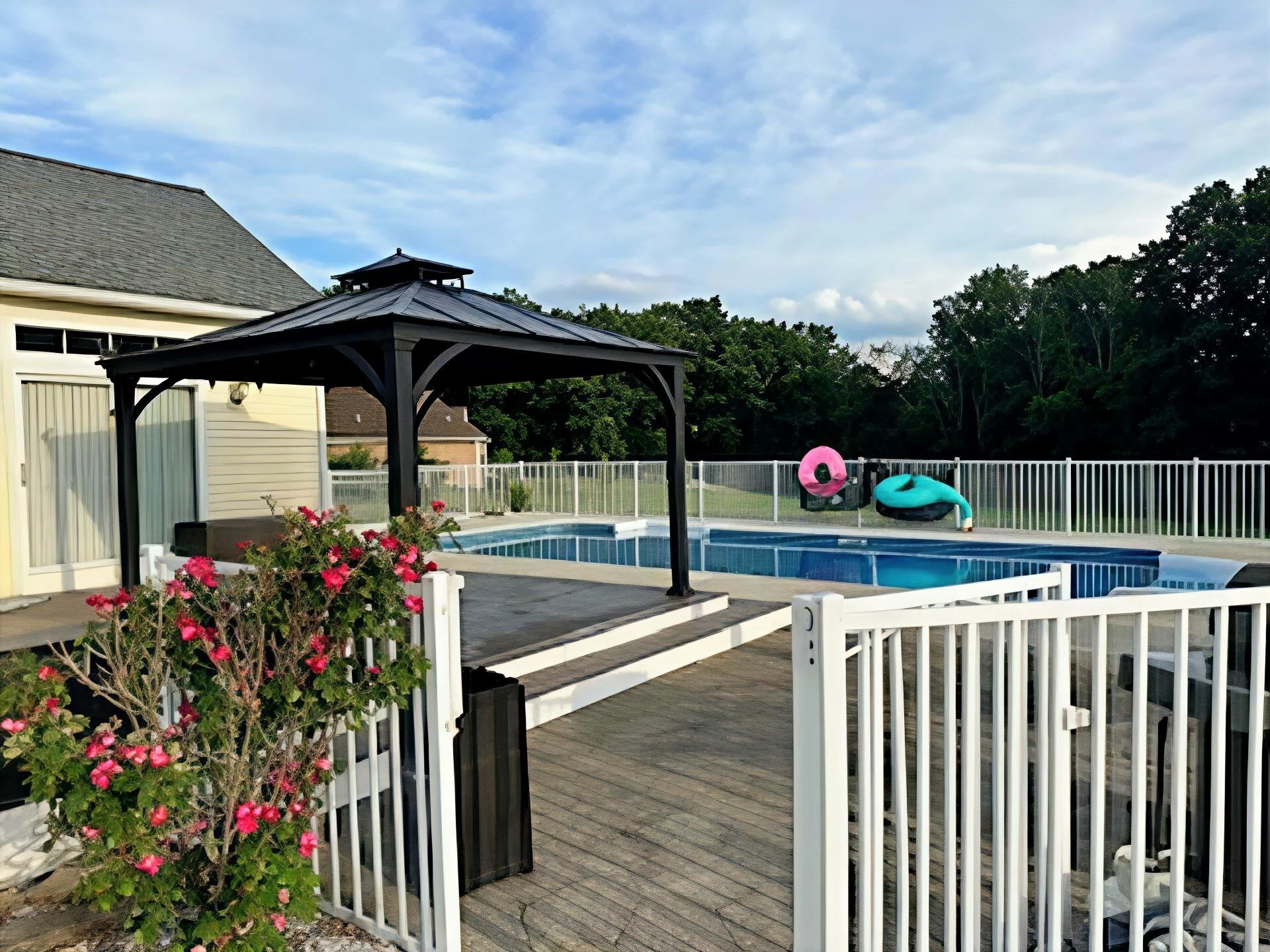 Residential backyard with swimming pool, gazebo, and two pool floats, pink donut and teal tube, surrounded by white fence and trees, under blue sky with clouds.