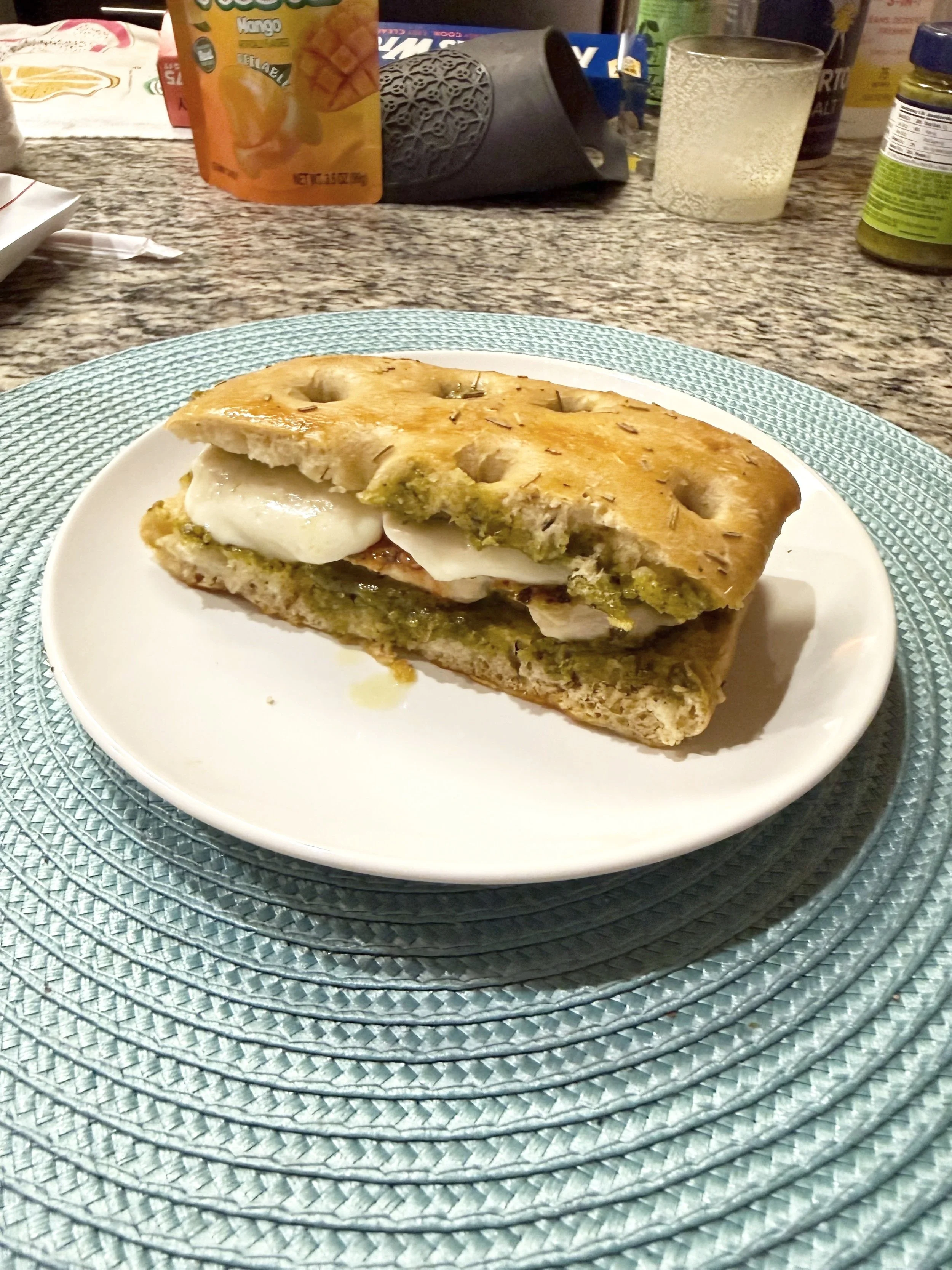 A sandwich on a white plate placed on a clear, textured placemat on a kitchen counter with various food items and beverages in the background.