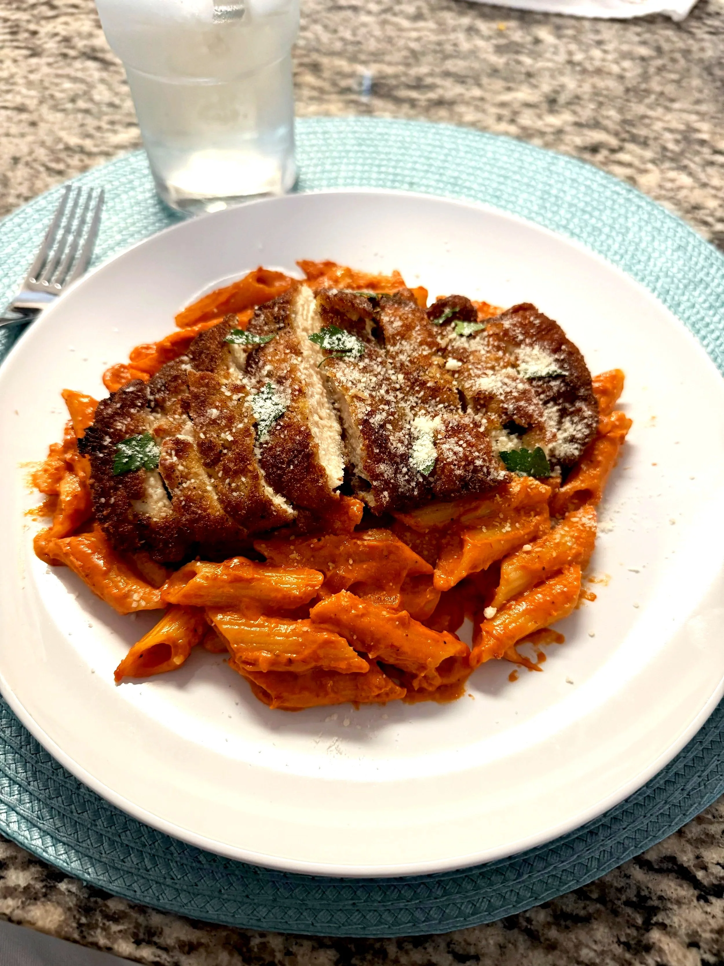 A plate of penne a la vodka topped with breaded and fried chicken cutlet, garnished with parsley, served on a white plate on a turquoise placemat on a granite table, with a glass of water in the background.