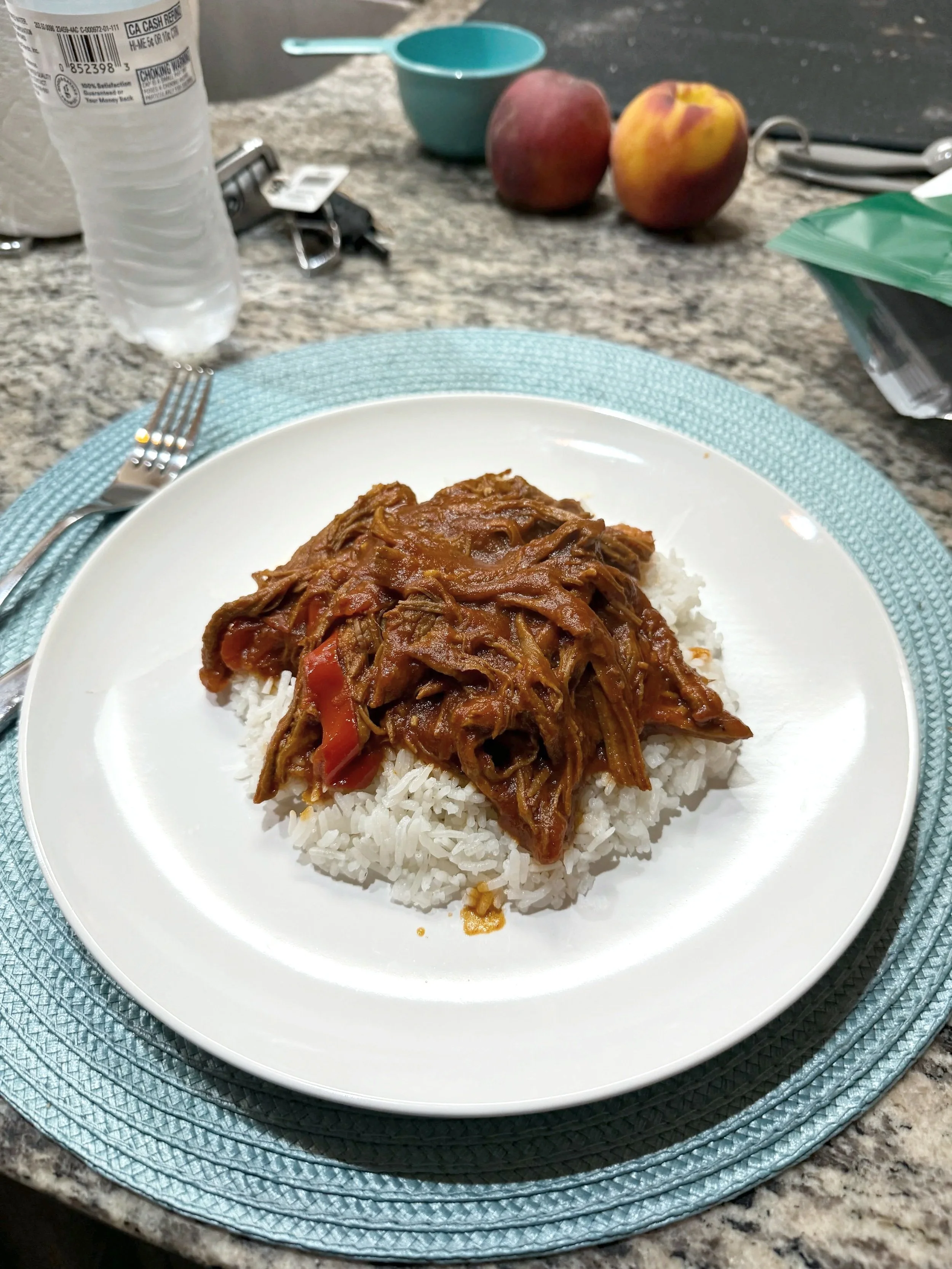 A plate of rice topped with shredded beef in sauce placed on a woven blue placemat on a granite countertop. In the background, two peaches, a water bottle, a small blue bowl, and miscellaneous items are visible.
