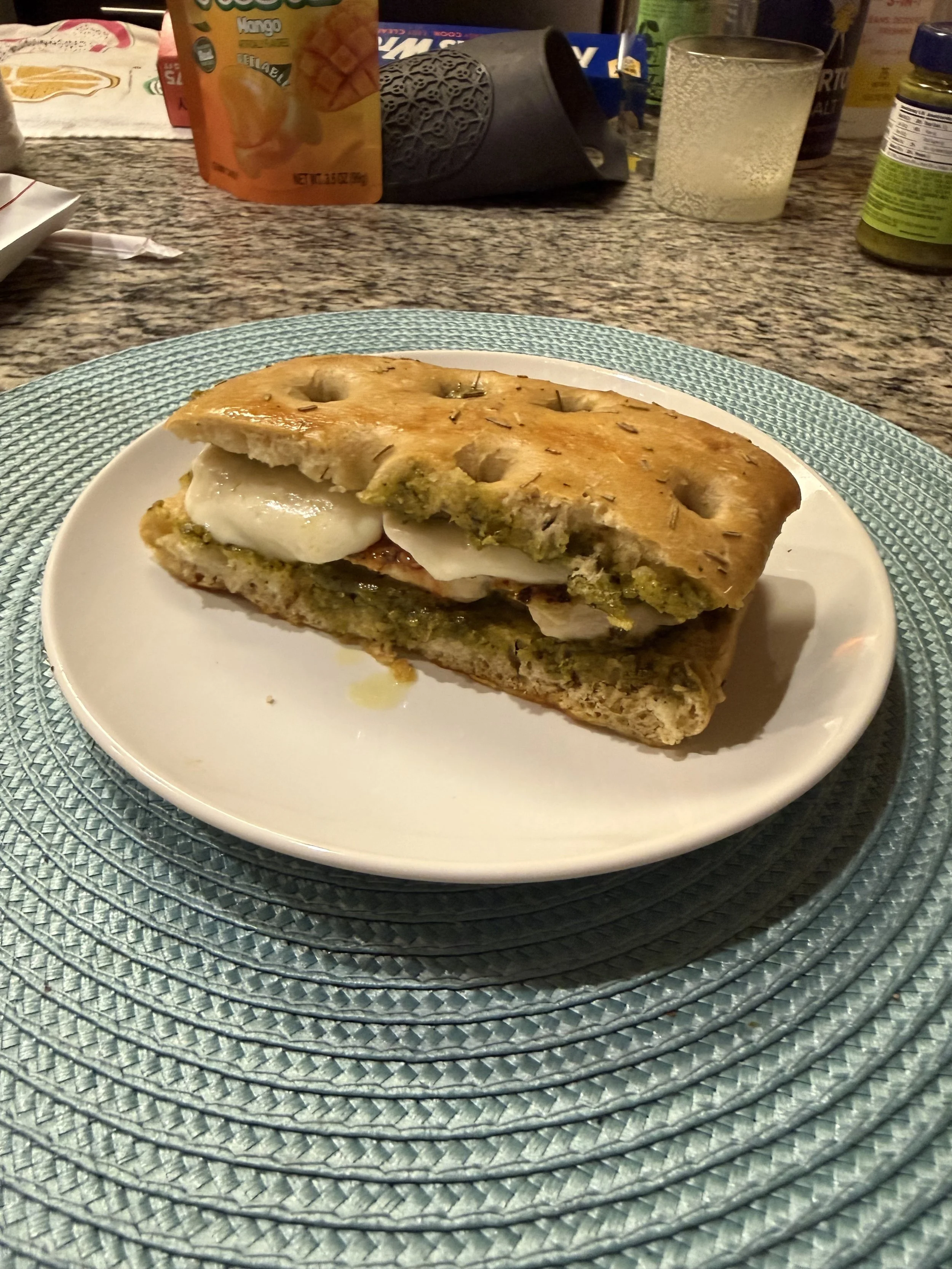 A sandwich with melted cheese and pesto sauce on a plate, placed on a blue textured placemat on a granite countertop, with various snack packages and a glass of beverage in the background.