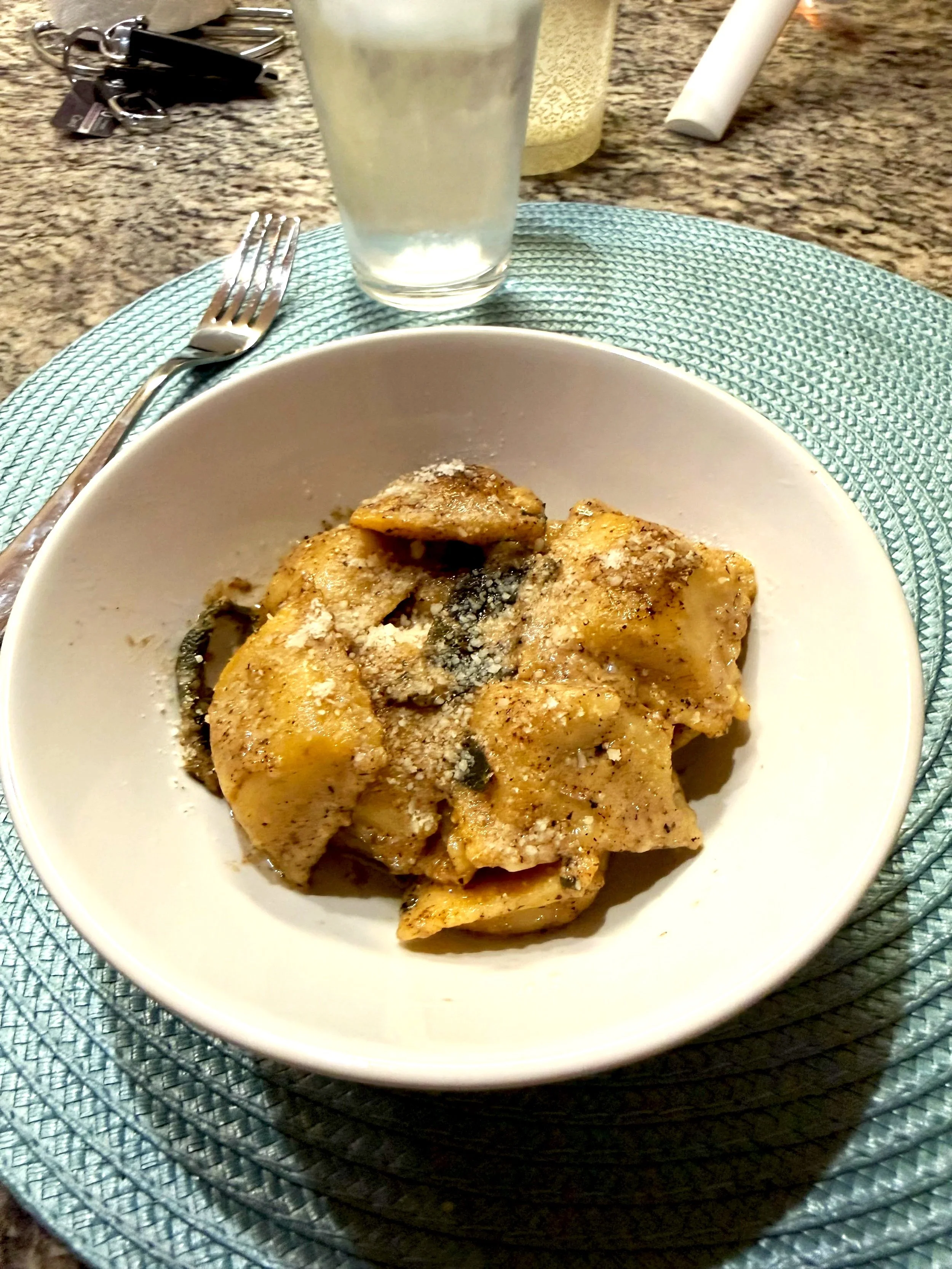 A bowl of brown butter sage butternut squash ravioli and grated cheese on top, placed on a turquoise placemat on a kitchen countertop with a fork to the left and a glass of water behind.