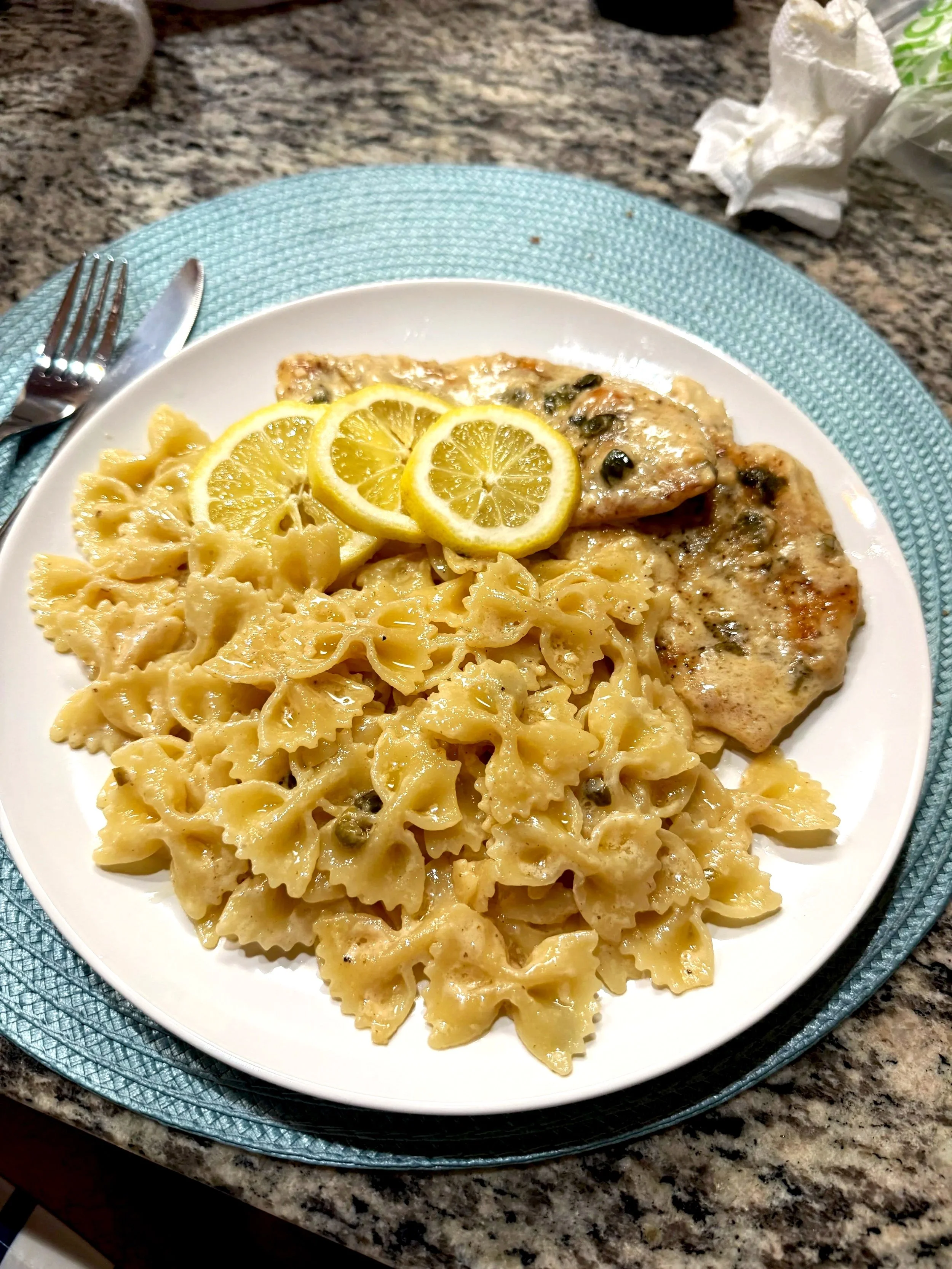 Plate of cooked bowtie pasta with creamy piccata sauce, topped with three lemon slices, and a piece of grilled chicken with herbs and lemon slices on top.