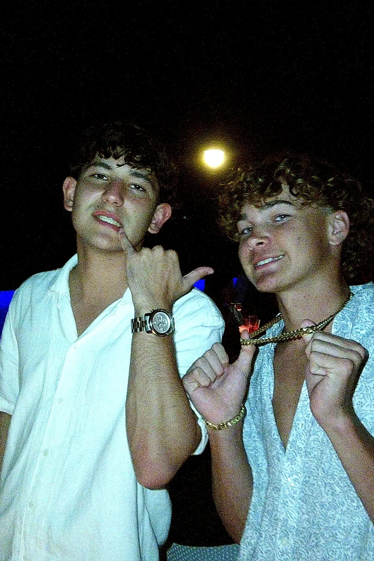 Two young men with curly hair posing with one showing a gold chain, at night with a bright moon overhead.