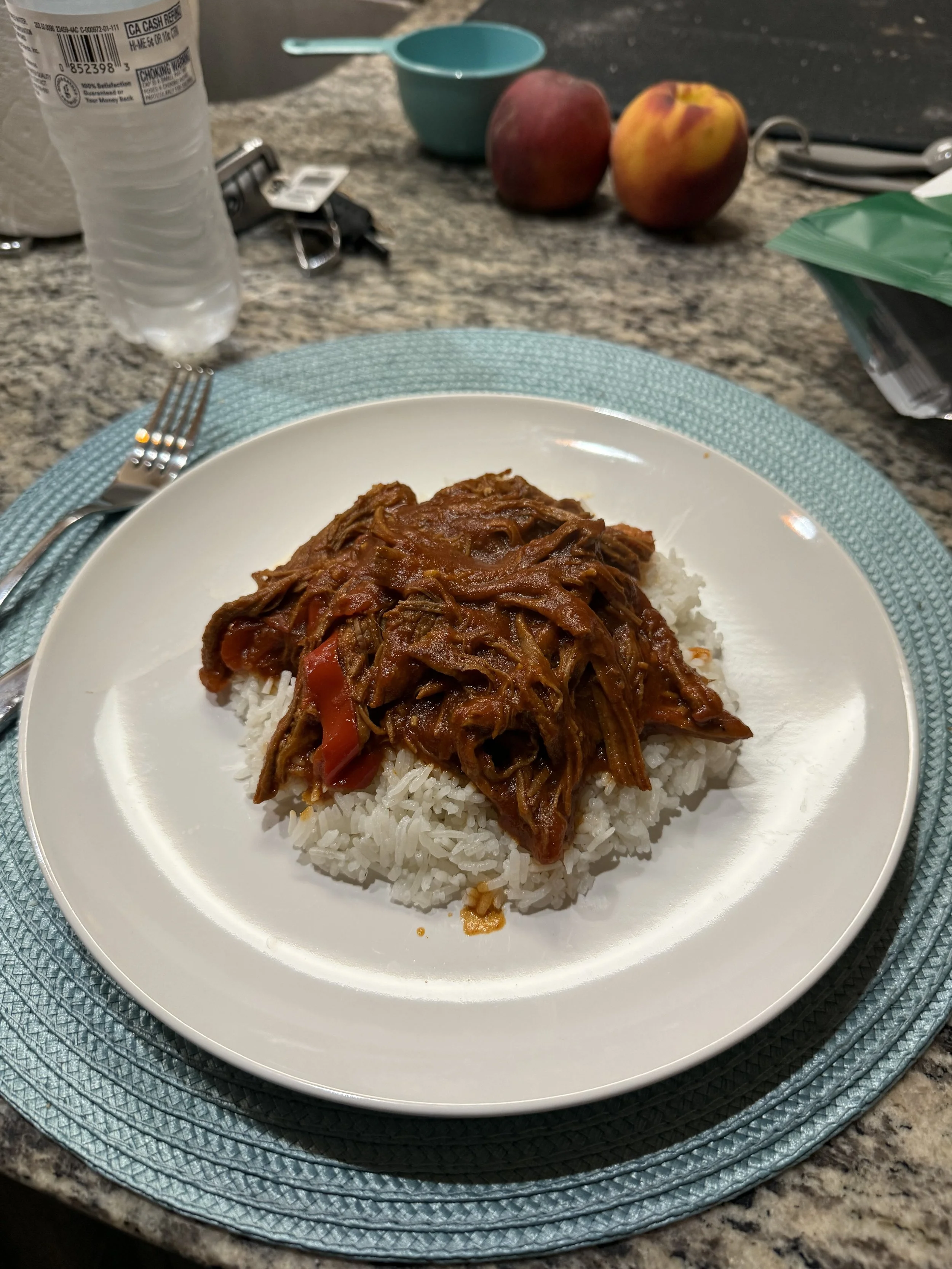 A plate of cooked shredded beef in sauce served over white rice on a blue placemat. In the background, there are two peaches, a plastic measuring cup, a clear bottle of water, and other kitchen items on a granite countertop.