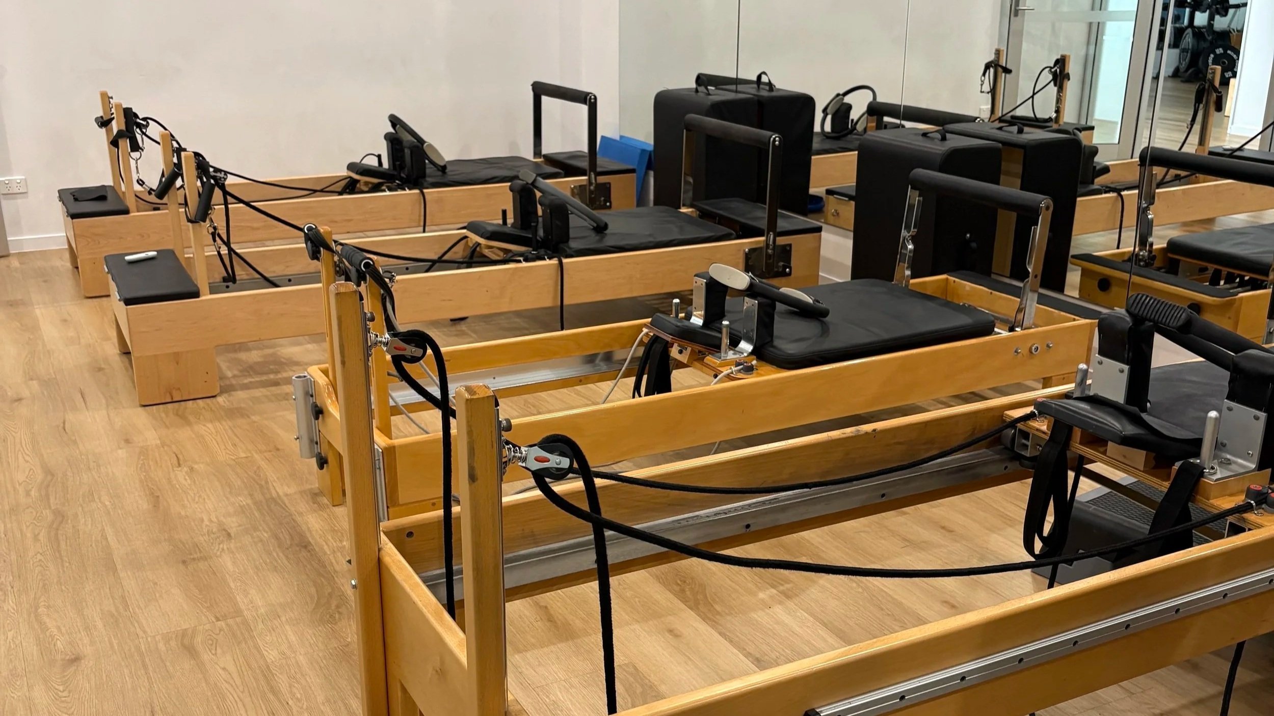 A Pilates reformer machine with wooden frame, black padded surfaces, and resistance straps in a fitness studio.