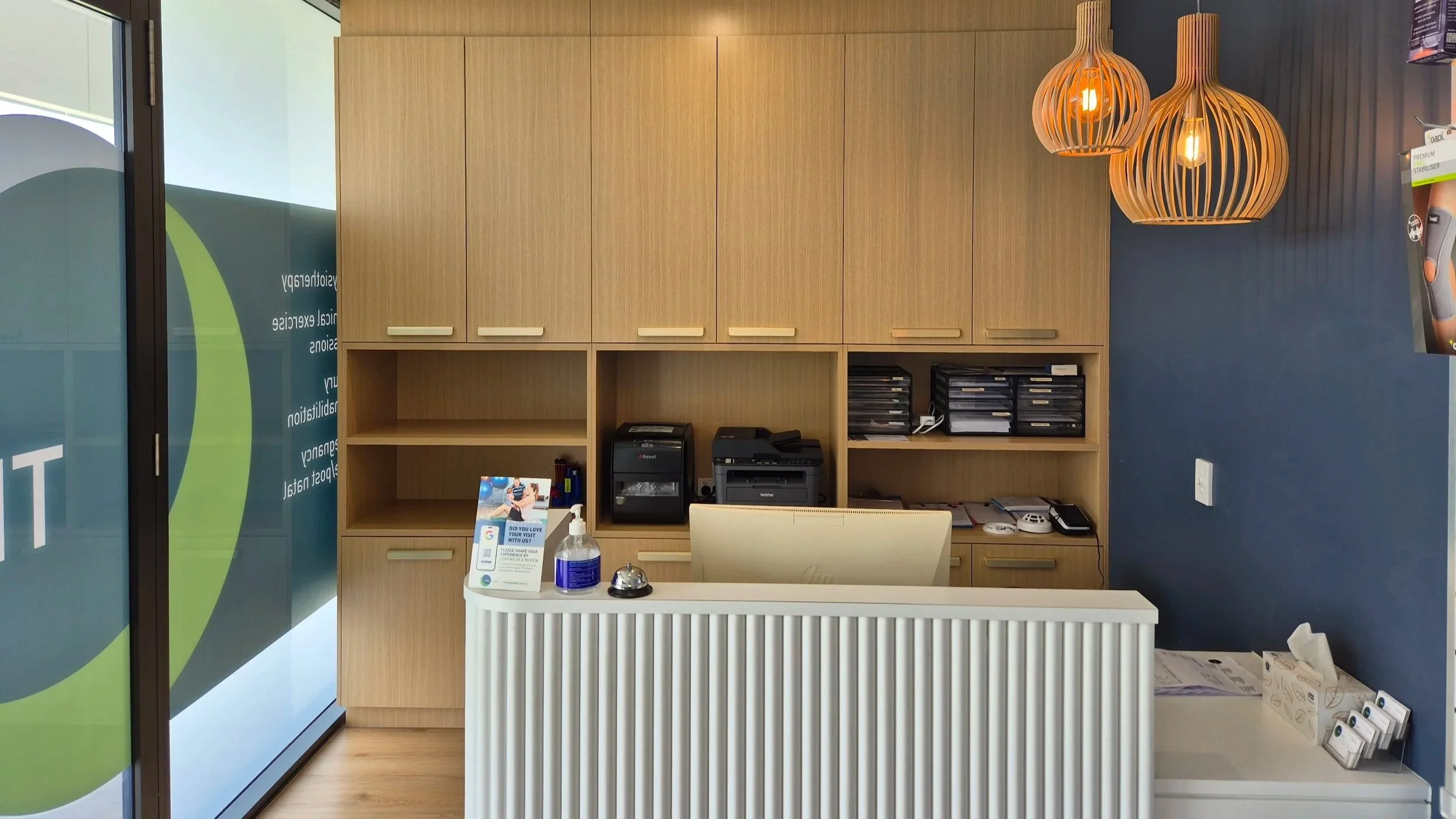 TPS Bayside Capalaba office reception area with a white desk, wooden cabinets, orange hanging pendant lights, a computer monitor, and a blue wall.