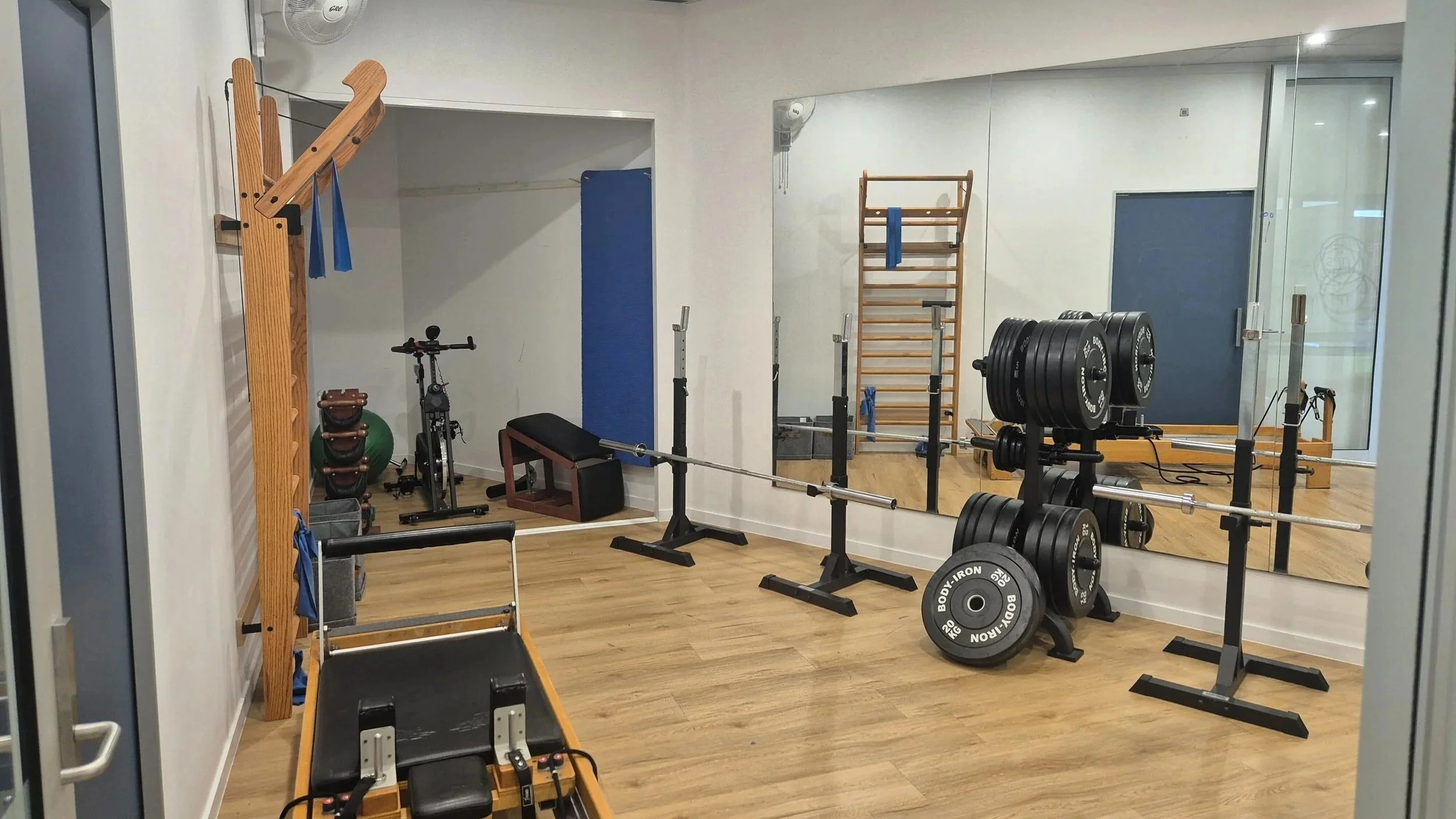 A physiotherapy gym room with wooden flooring, containing various fitness equipment such as weights, an exercise bike, a bench, and wall-mounted pull-up bars. There is a large mirror on the wall reflecting part of the gym and a doorway on the right.