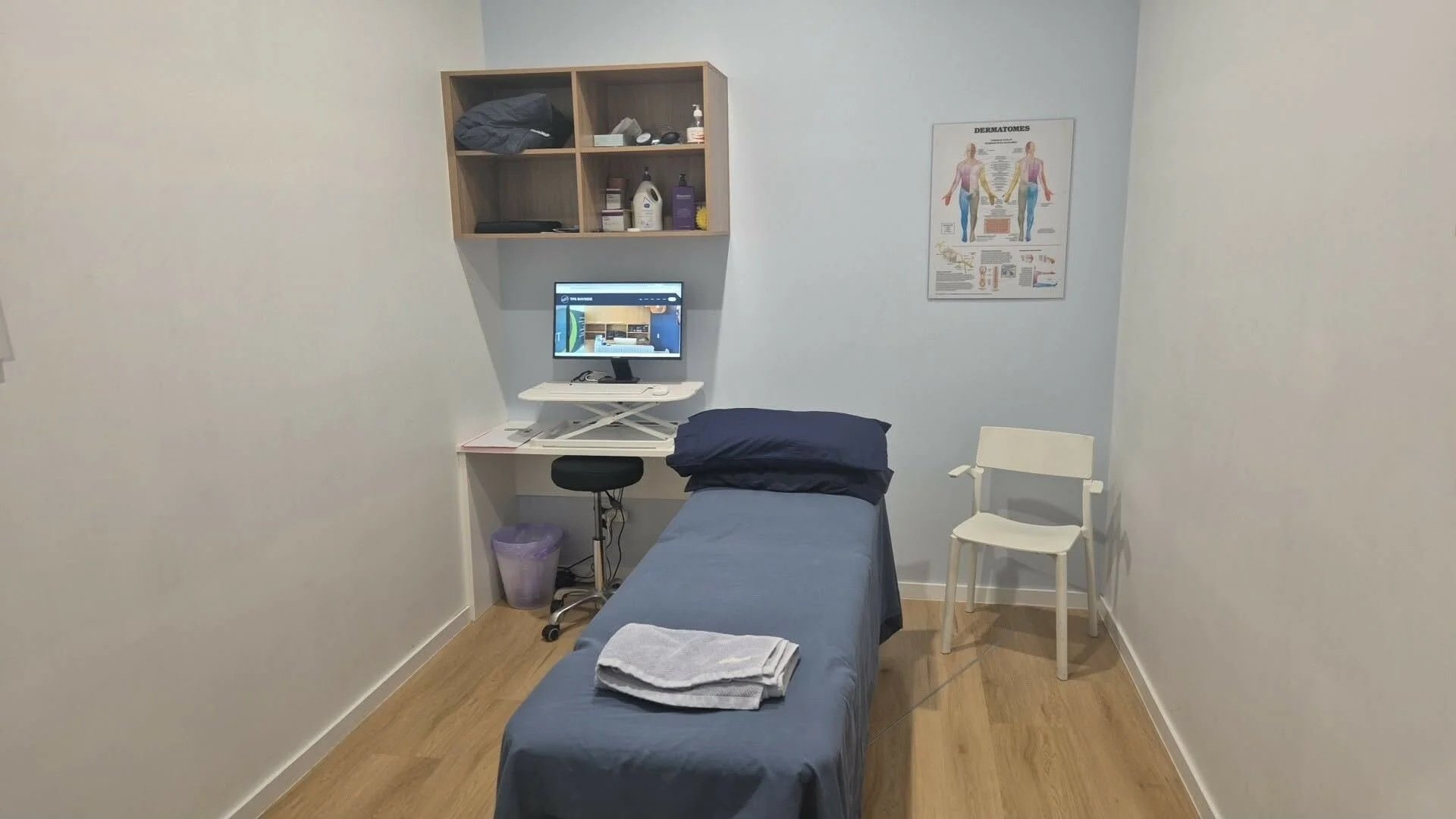 TPS Bayside Victoria Point physiotherapy examination room with a bed, pillow, folded towels, computer on a rolling desk, a chair, cabinet with medical supplies, a white chair, and an anatomy poster on the wall.