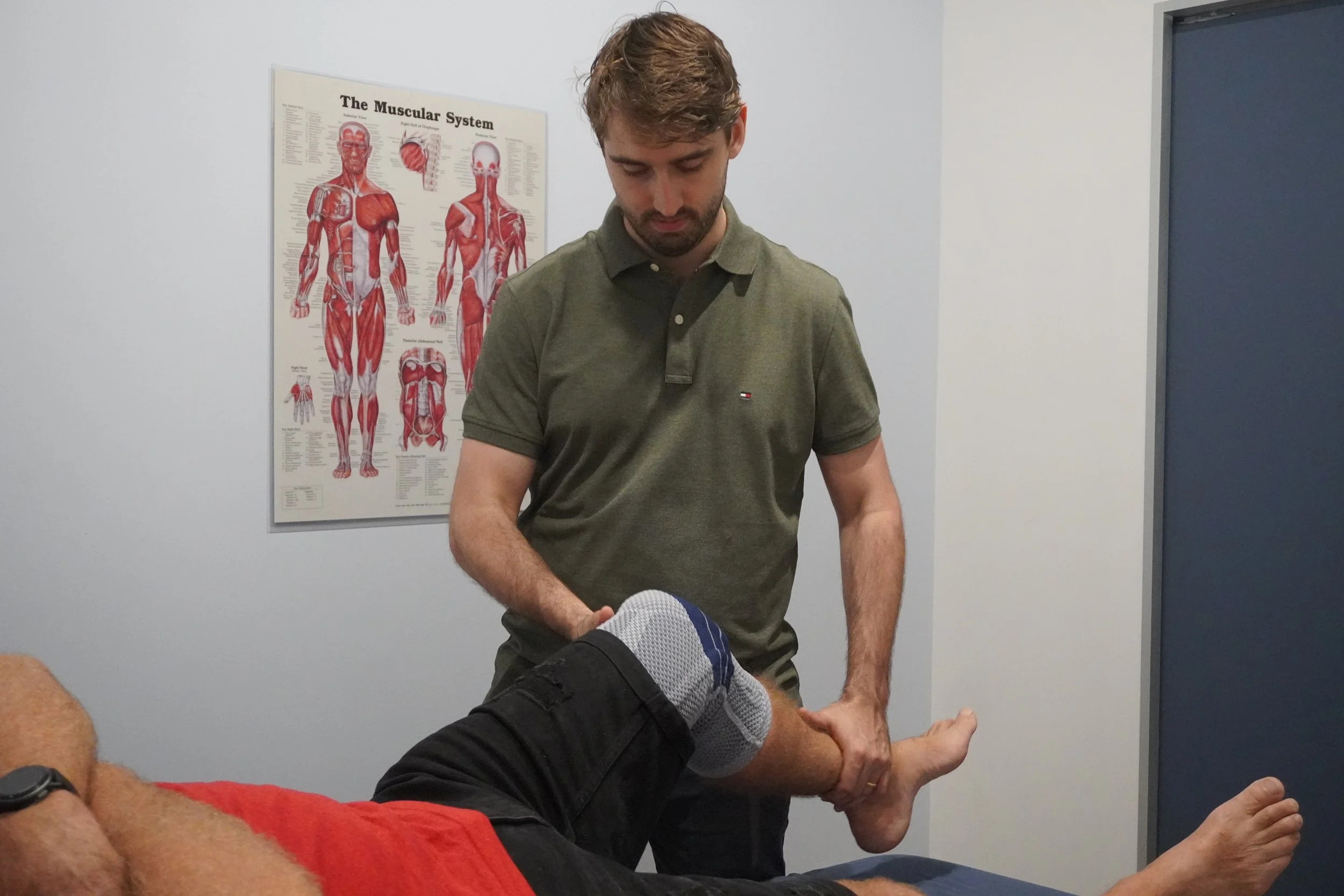 A man in a green polo shirt examining another man's knee in a physiotherapy clinic with a diagram of the muscular system on the wall.