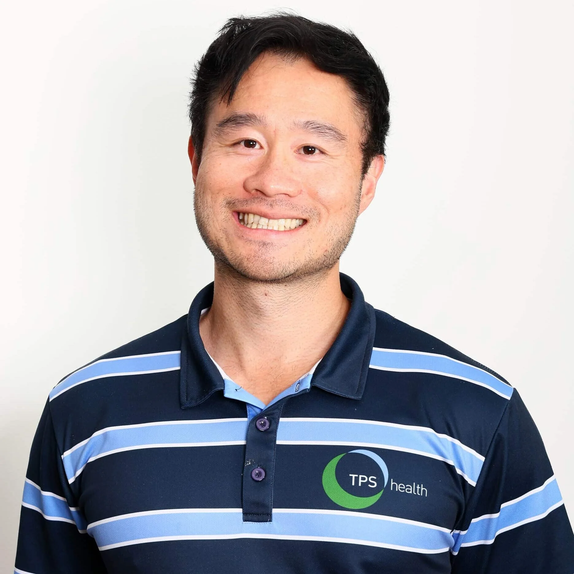 Smiling male physiotherapist wearing a TPS Bayside Capalaba and Victoria Point navy blue and light blue striped polo shirt standing against a plain white background.