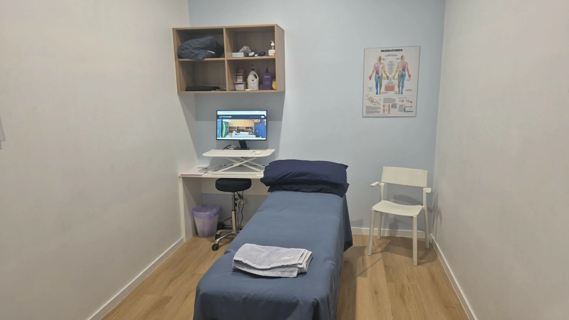 TPS Bayside Victoria Point physiotherapy examination room with a bed, pillow, folded towels, computer on a rolling desk, a chair, cabinet with medical supplies, a white chair, and an anatomy poster on the wall.