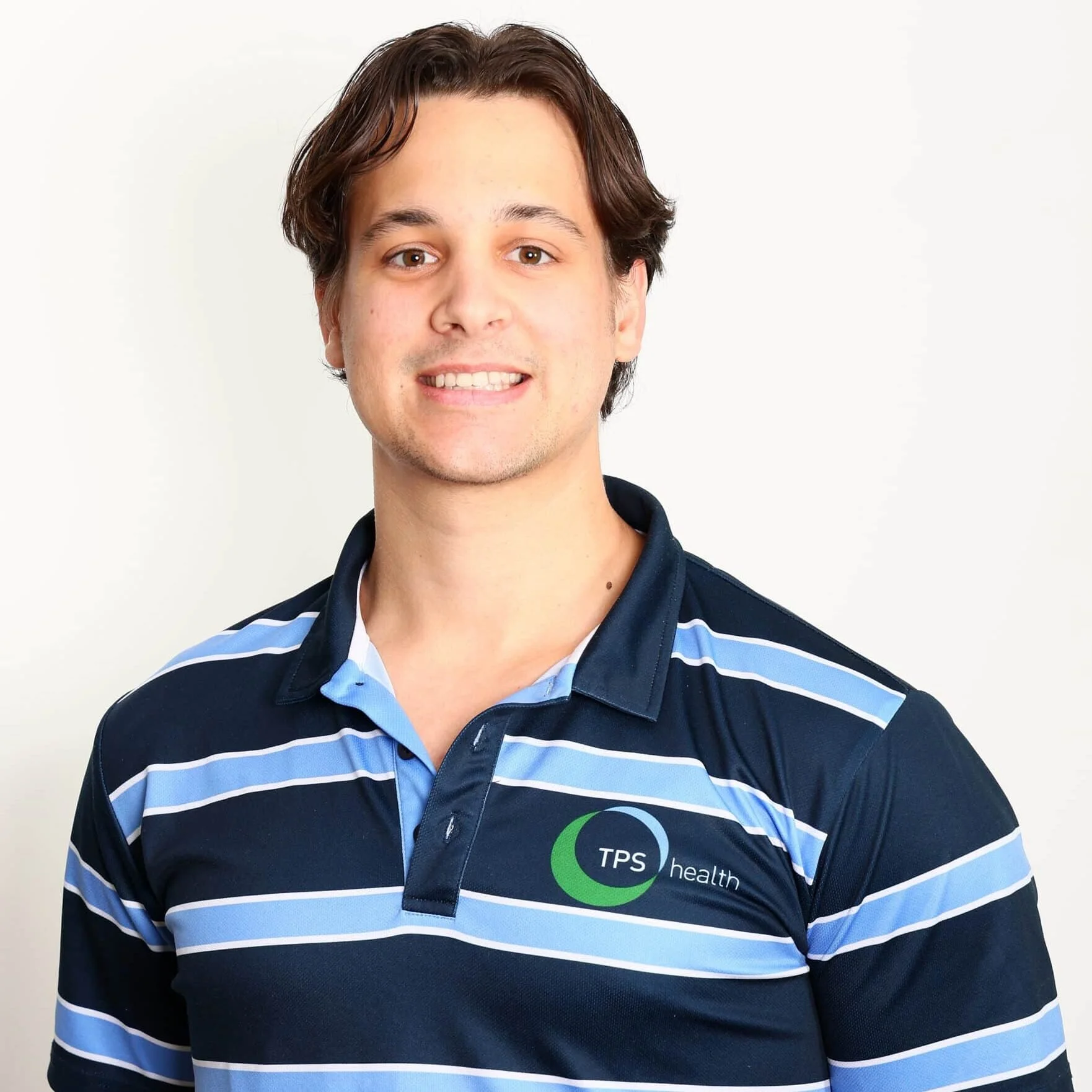 Smiling male physiotherapist wearing a TPS Bayside Capalaba and Victoria Point navy blue and light blue striped polo shirt standing against a plain white background.