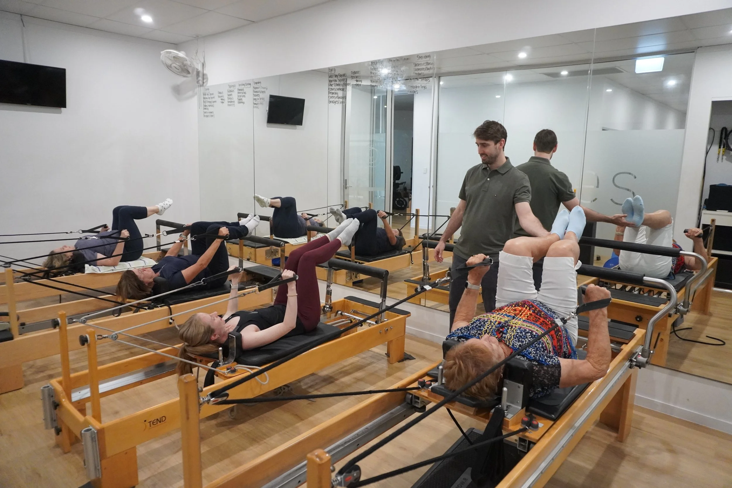 People participating in a Pilates reformer clinical exercise class in a physiotheraphy studio, led by a physiotherapist, with mirror wall and whiteboard detailing exercises in the background.