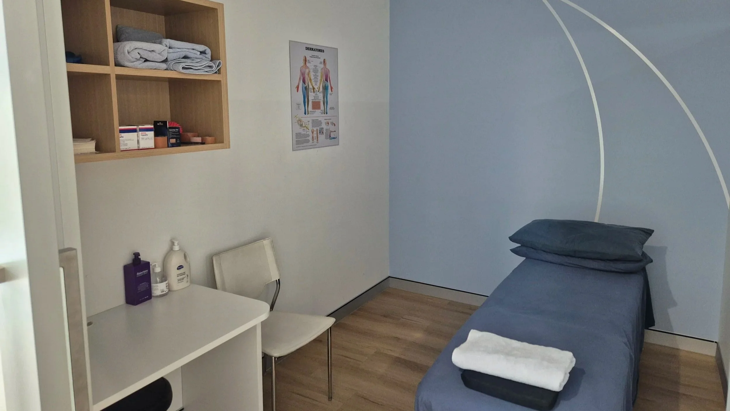 TPS Bayside Capalaba physiotherapy examination room with a bed, pillow, folded towels, computer on a rolling desk, a chair, cabinet with medical supplies, a white chair, and an anatomy poster on the wall.