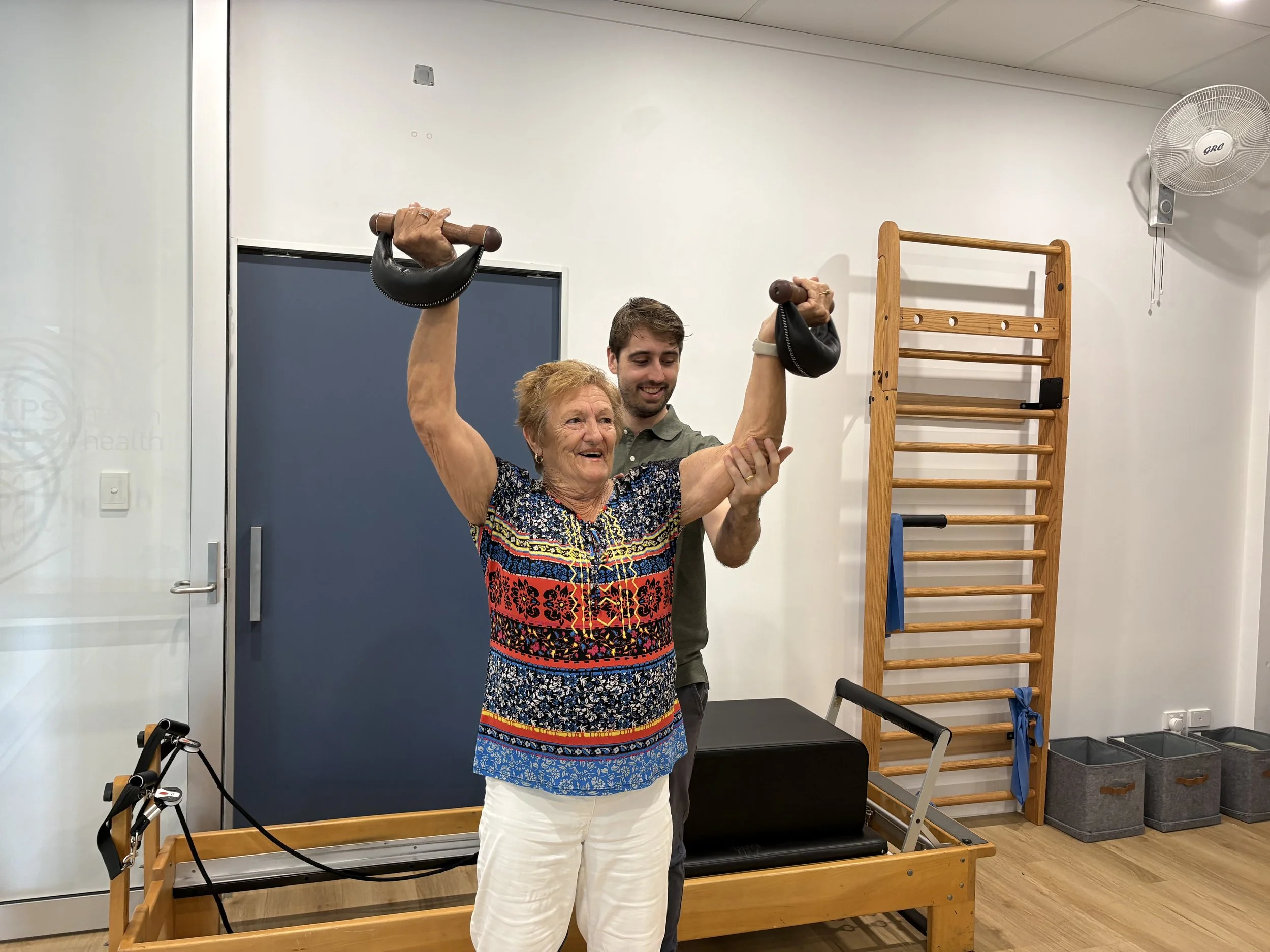 An elderly woman standing lifting hand weights in a physiotheraphy clinic during a bone building class. There is a wooden ladder-like exercise equipment and a table with exercise equipment nearby in the background.