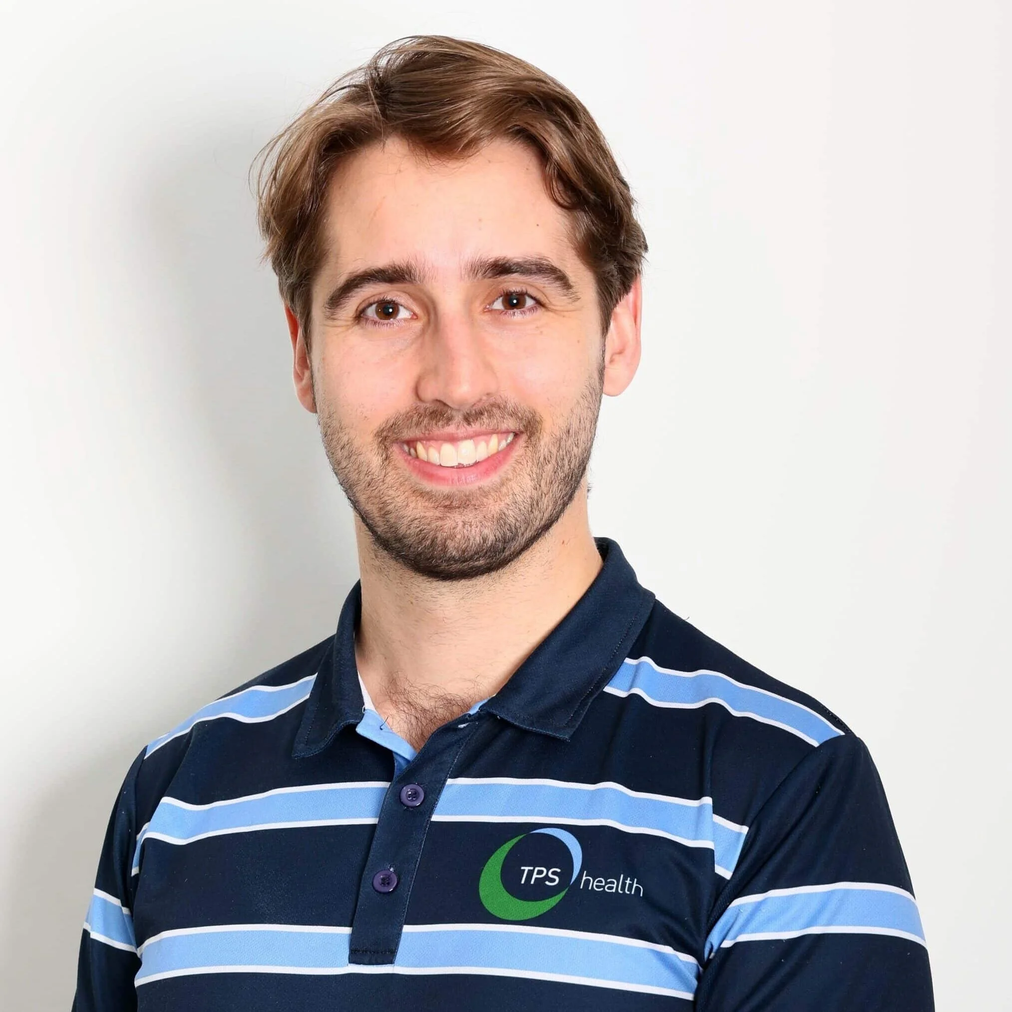 Smiling male physiotherapist wearing a TPS Bayside Capalaba and Victoria Point navy blue and light blue striped polo shirt standing against a plain white background.