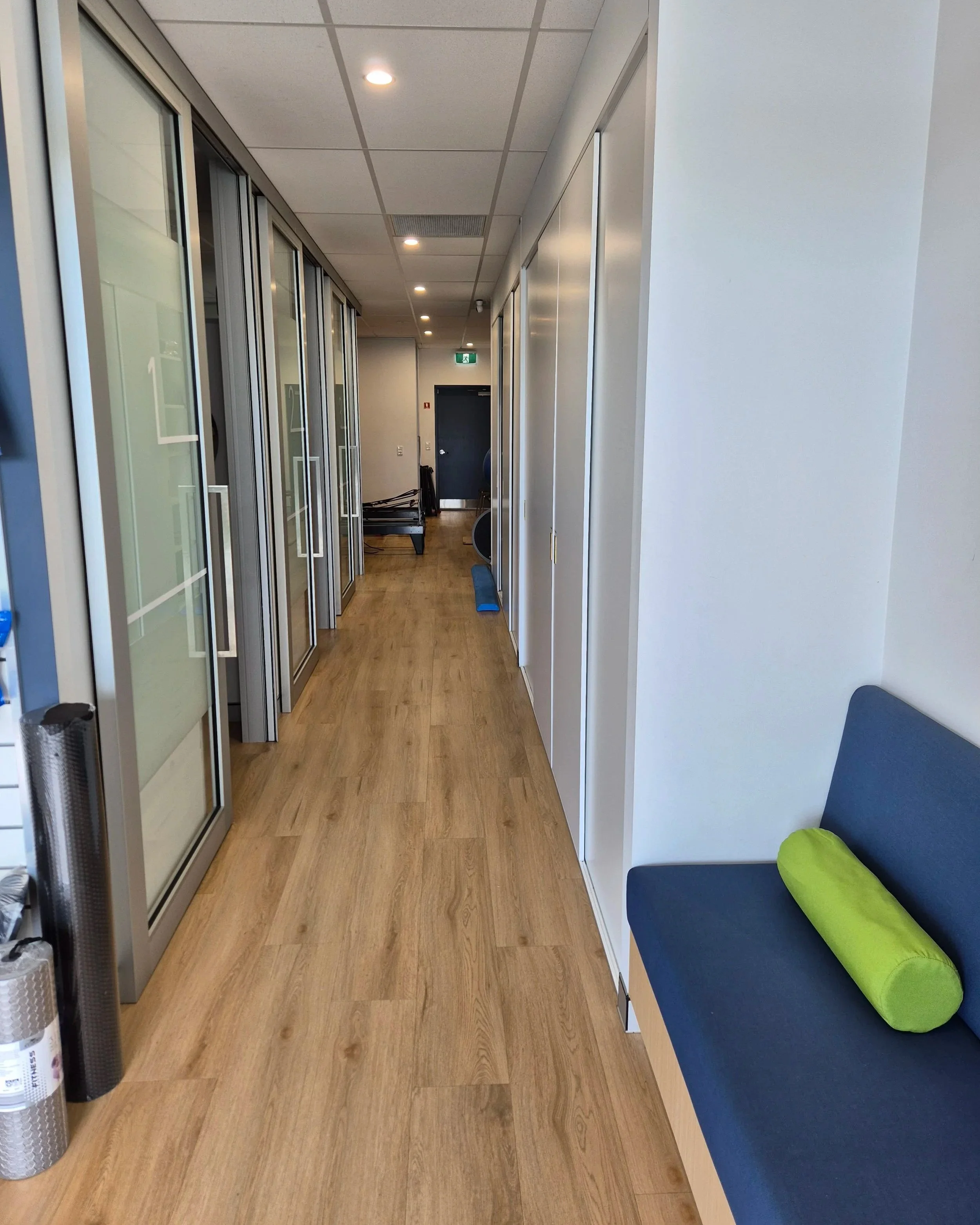 Corridor in TPS Bayside's Capalaba clinic with wooden flooring, glass partition walls to physiotherapy rooms on the left, and a seating area with a blue cushion and a green cushion on the right. The hallway leads to a gym with reformers.