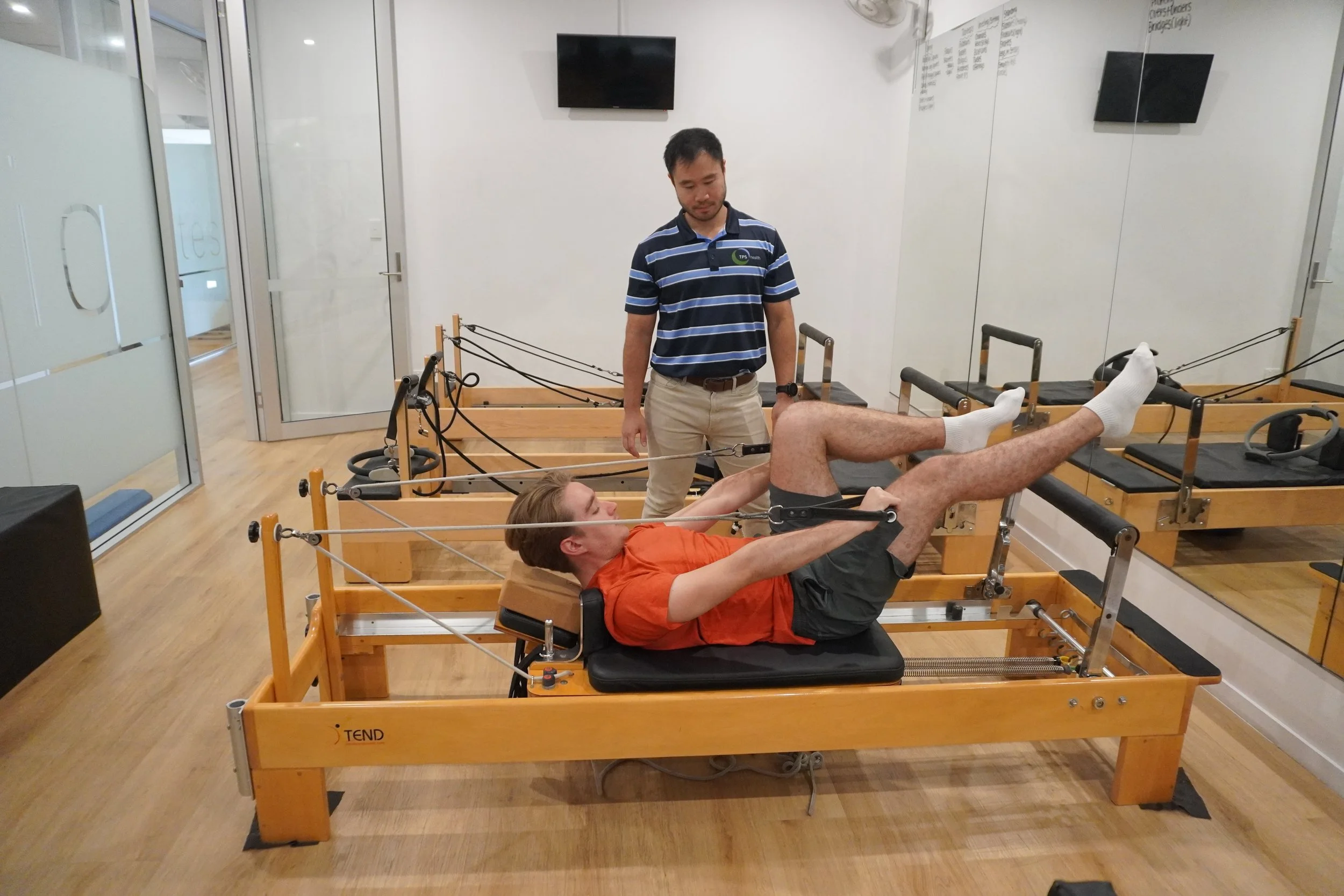 A man lying on a Pilates reformer machine exercising for injury rehabilitation with the assistance of a physiotherapist standing next to him in a gym.