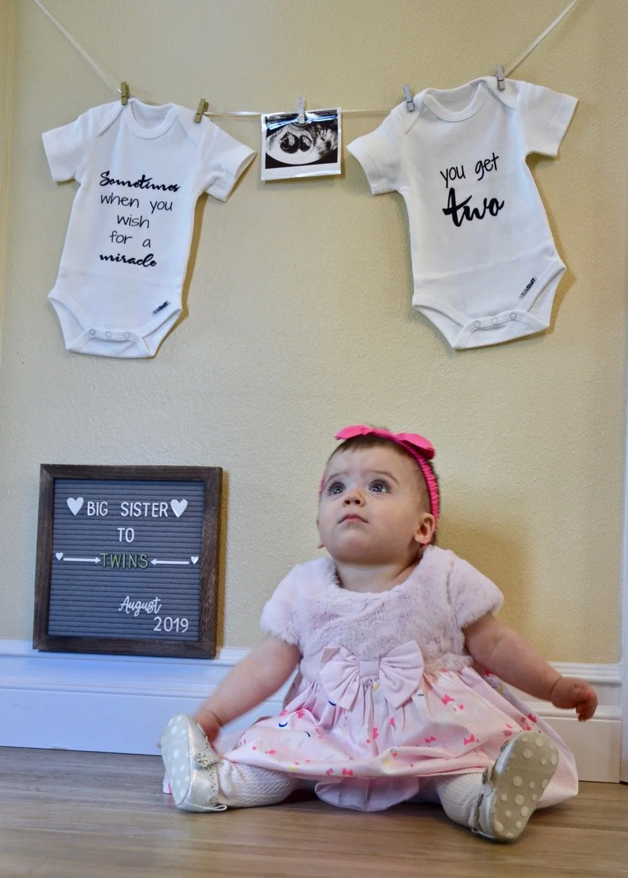 One year old gazing up at two onesie’s in the announcement of her twin brothers.