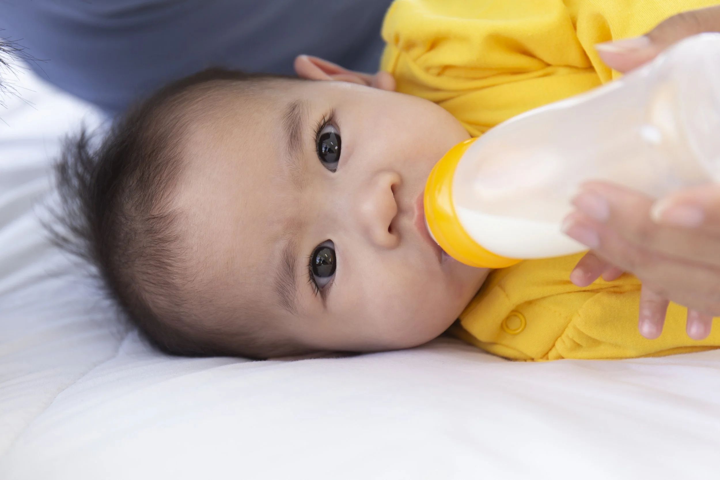 Close-up of a young child lying on a white surface, drinking milk from a bottle, wearing a yellow garment.