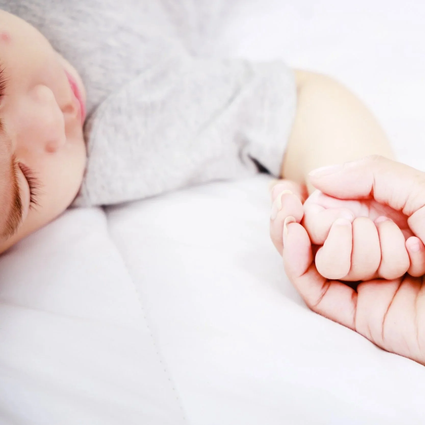 Close-up of a sleeping baby's face, with a hand gently holding the baby's tiny fingers. The baby is lying on a soft, light-colored surface and is dressed in a light gray top. The scene conveys a sense of calmness and tenderness.