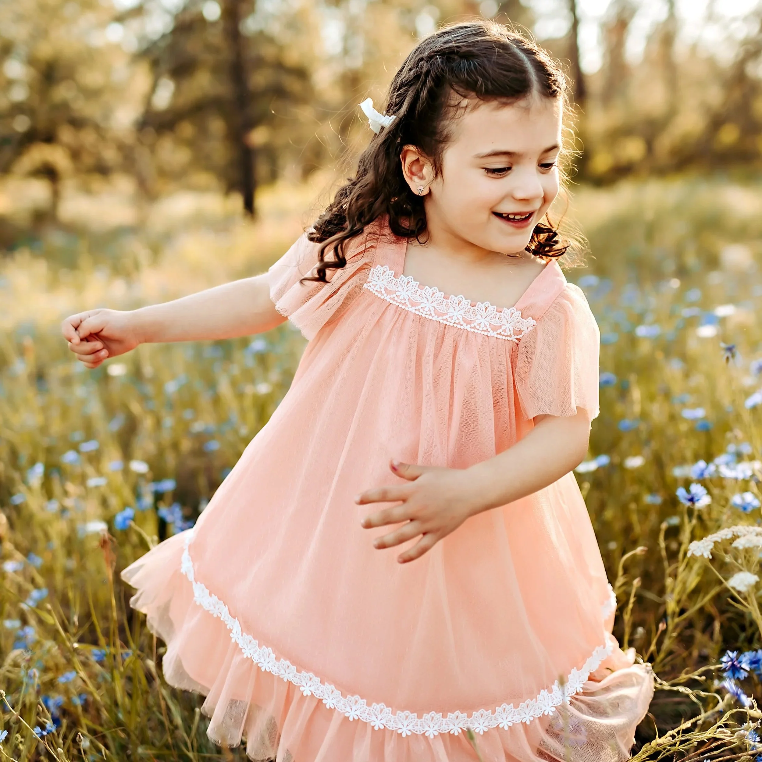 A young girl with curly dark hair in a braid, wearing a pastel pink dress with white lace accents, is outdoors in a field with tall grass and wildflowers, smiling and looking down.