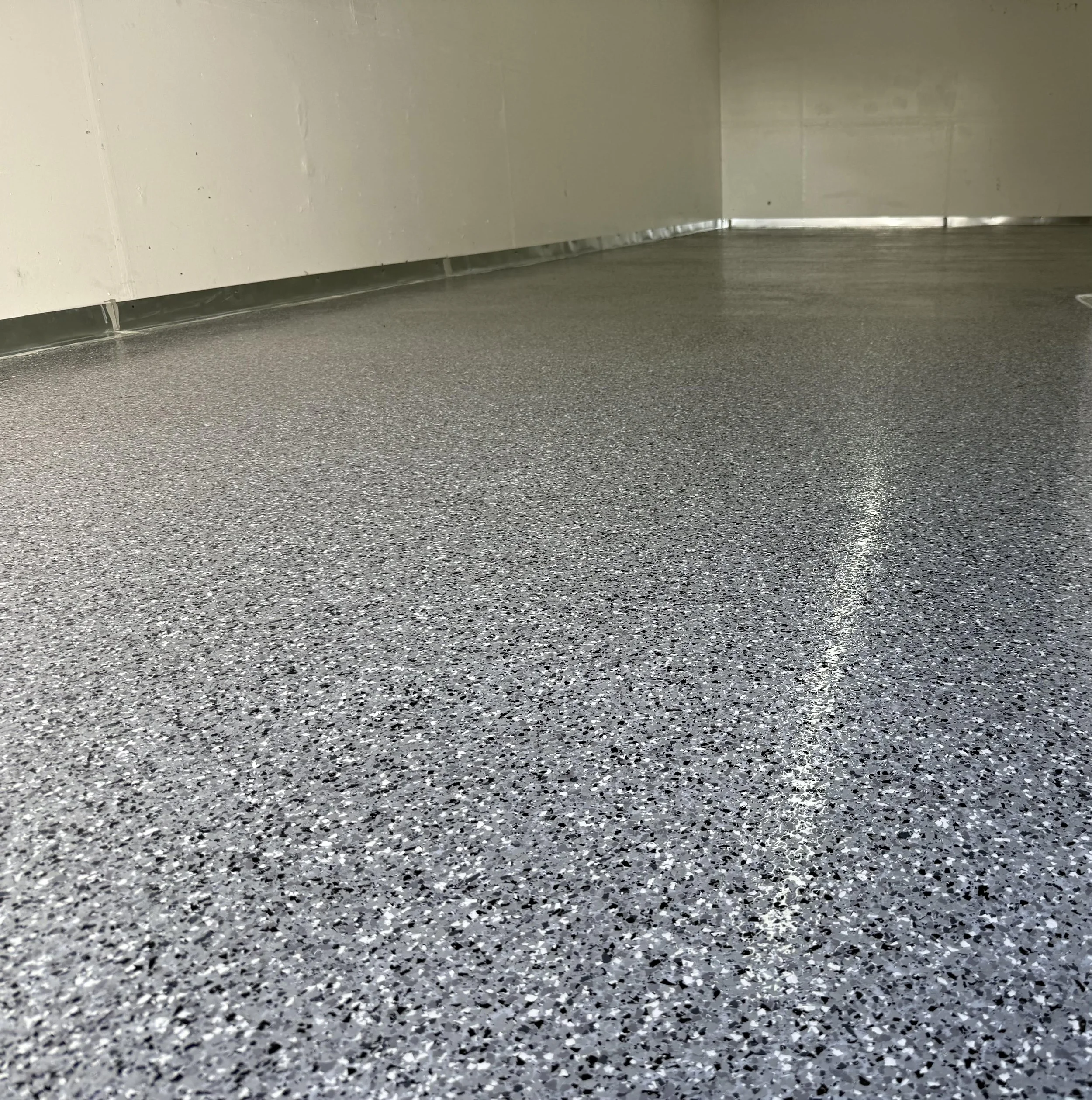 Close-up of gray speckled flooring in an indoor space with a beige wall and metallic baseboard.